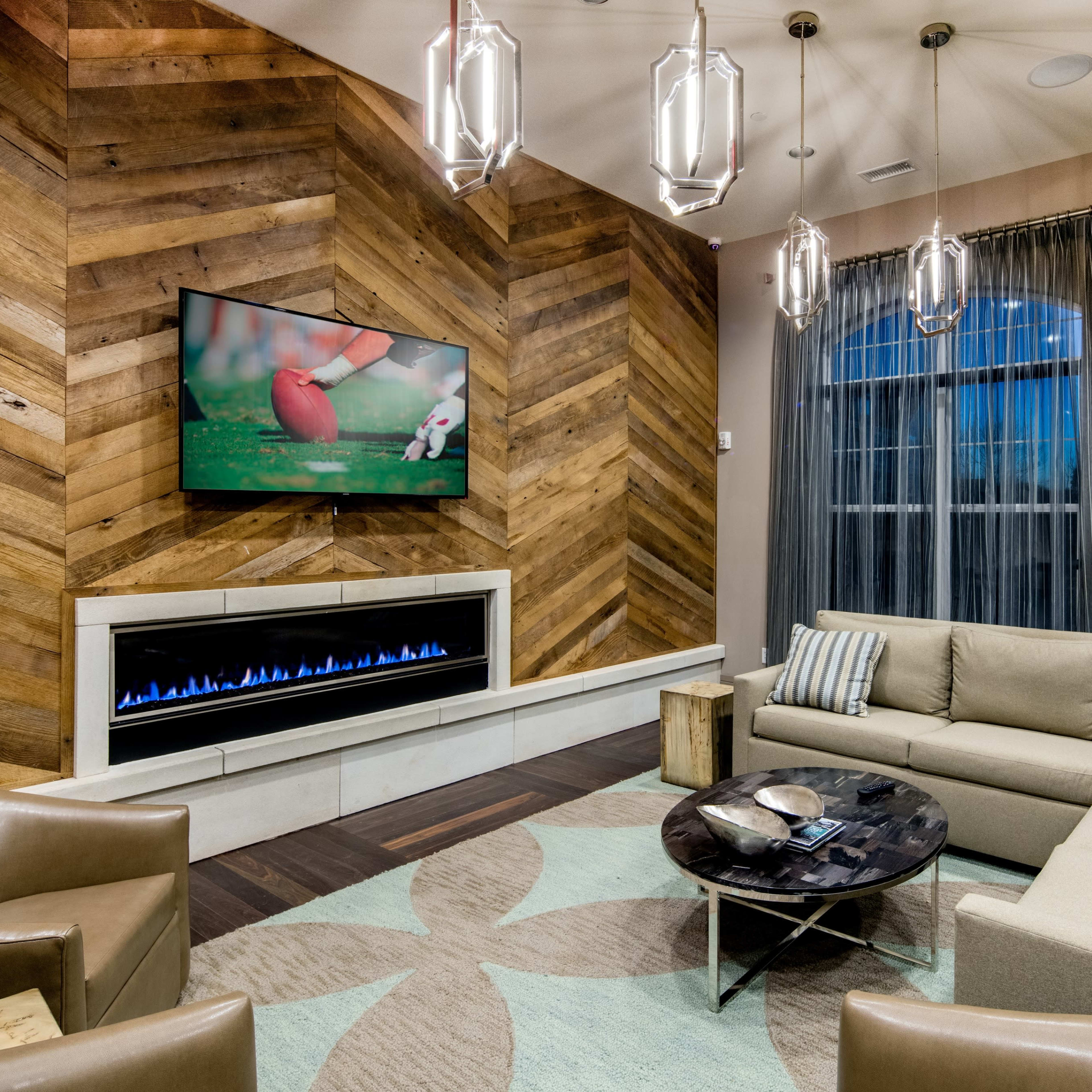 A modern living room featuring a sectional sofa, armchairs, a round coffee table, a wall-mounted television, and a fireplace beneath the TV, all set against a backdrop of wood-paneled walls.