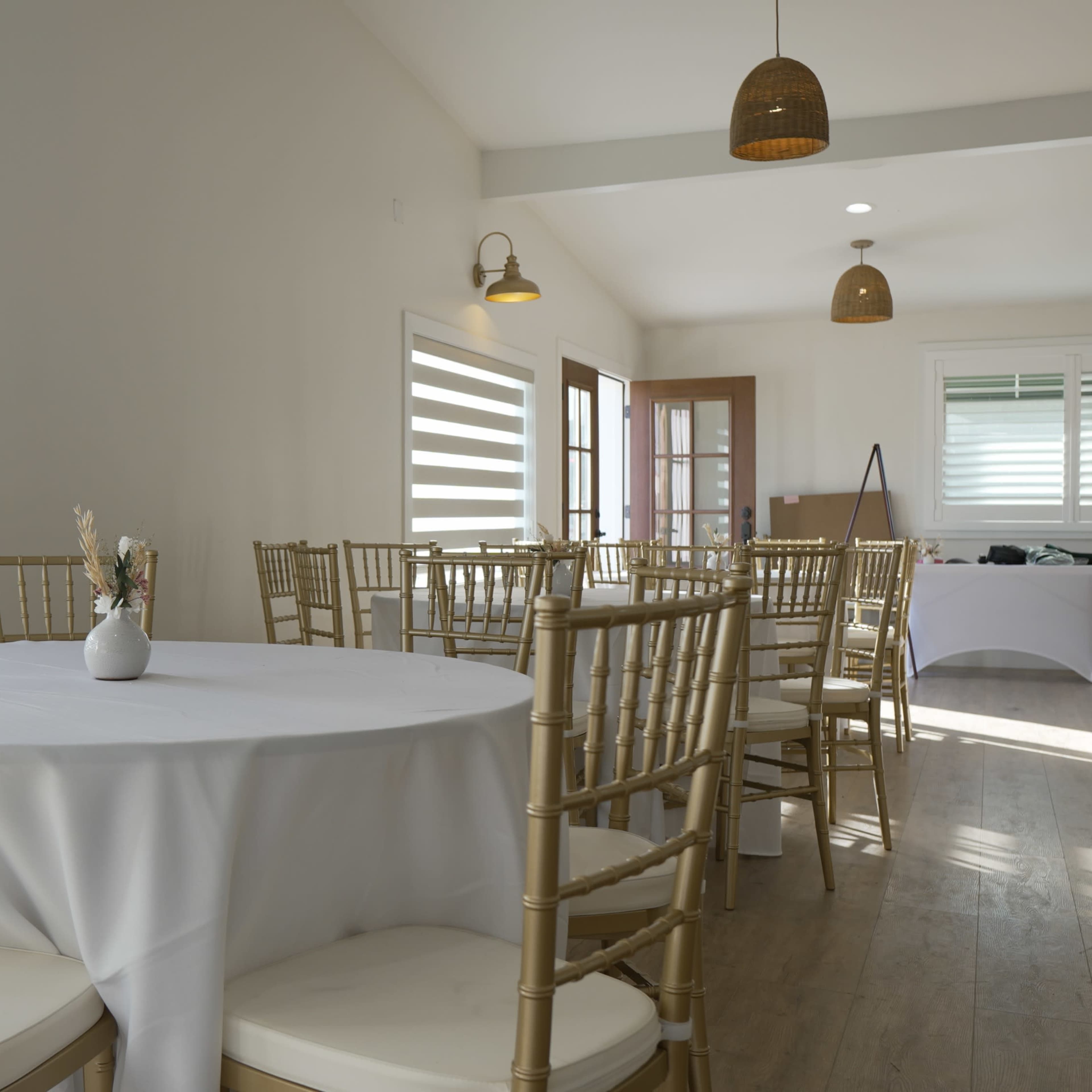 A bright, minimalist room features round tables with white tablecloths and gold chiavari chairs, illuminated by natural light through striped windows.