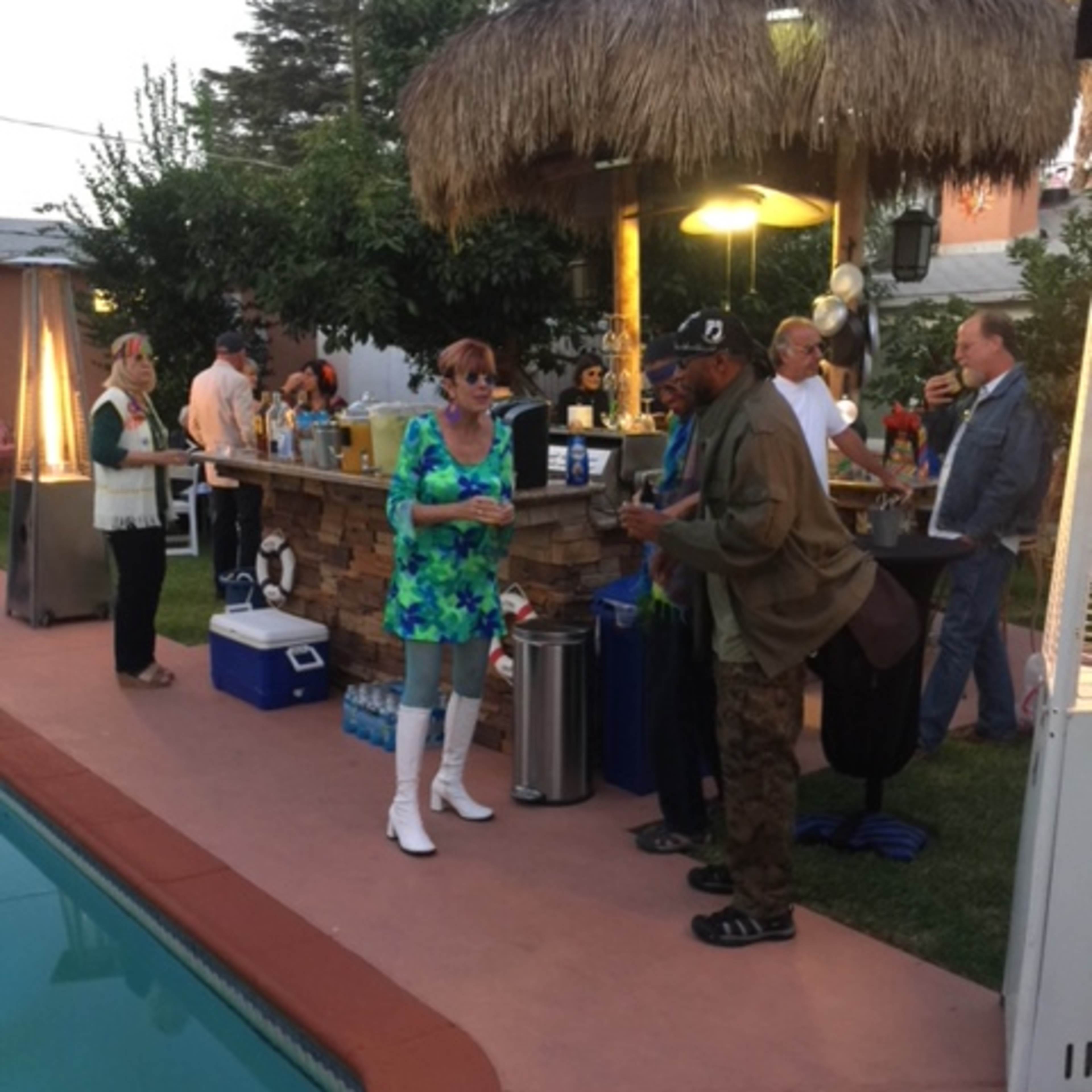 A group of people socializes around a poolside bar with a thatched roof at an evening gathering.