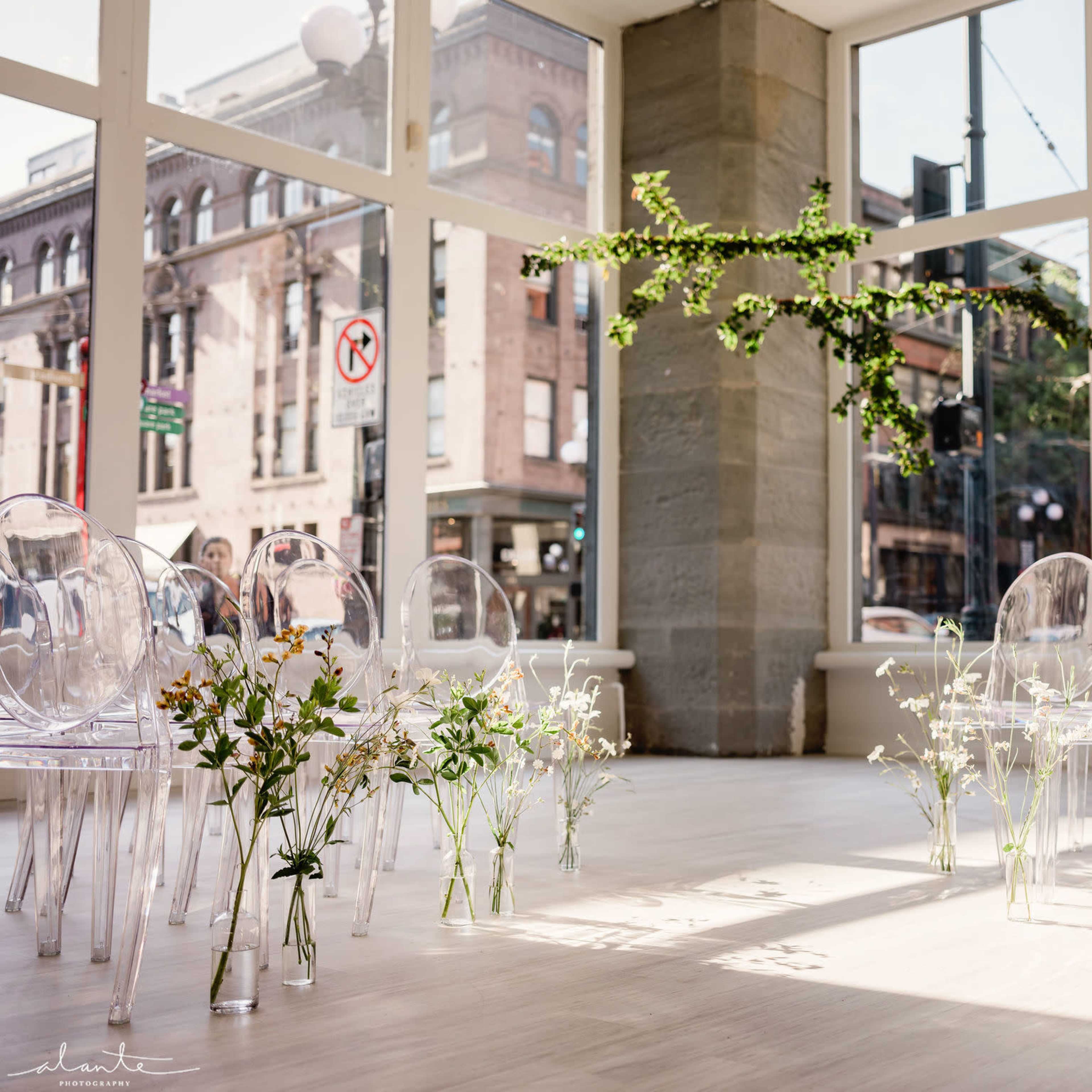 A brightly lit indoor space with clear chairs arranged in rows, surrounded by small floral arrangements on the floor and a hanging greenery installation above.