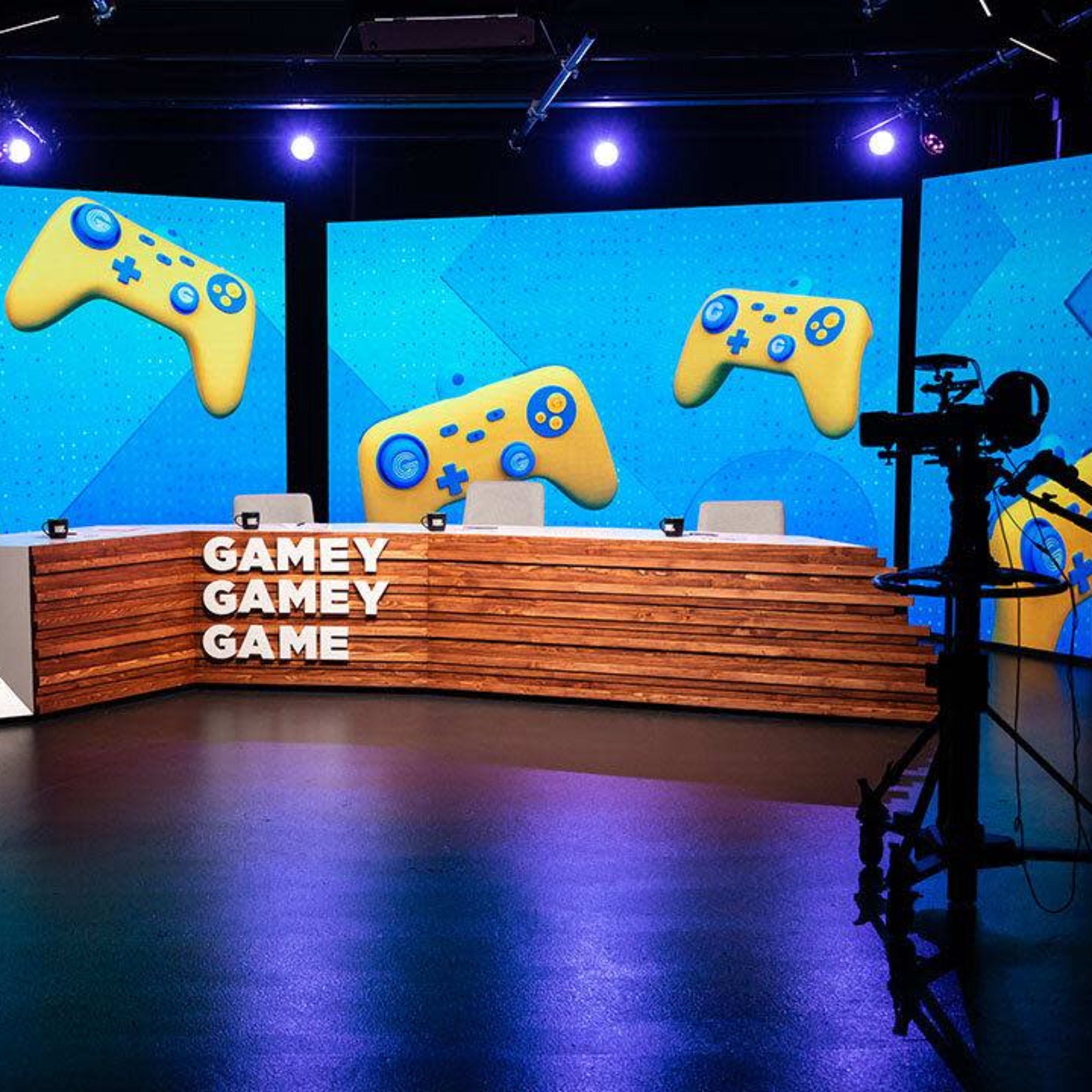 A television studio set features a wooden desk with the words "GAMEY GAMEY GAME" and bright blue backgrounds displaying yellow gaming controllers.