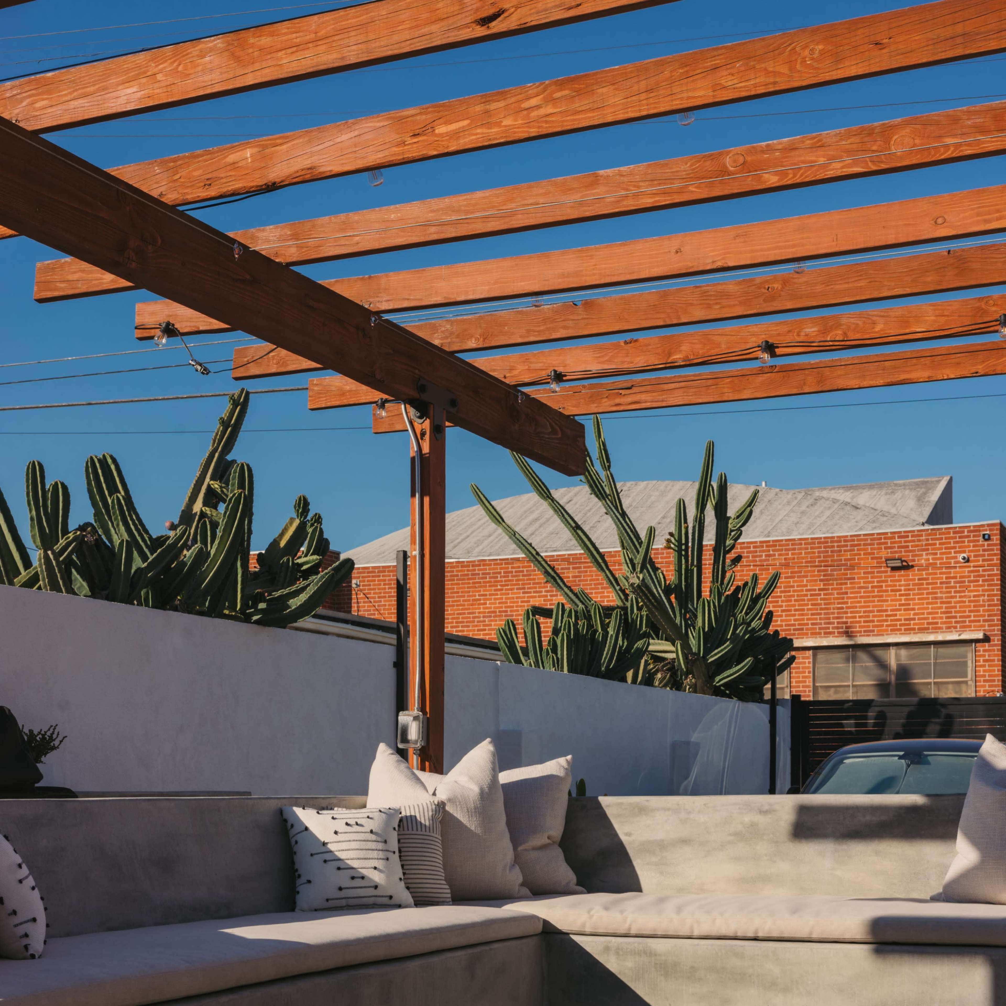 A modern outdoor seating area with a concrete bench, decorative pillows, and a circular fire pit under a wooden pergola, surrounded by cacti.