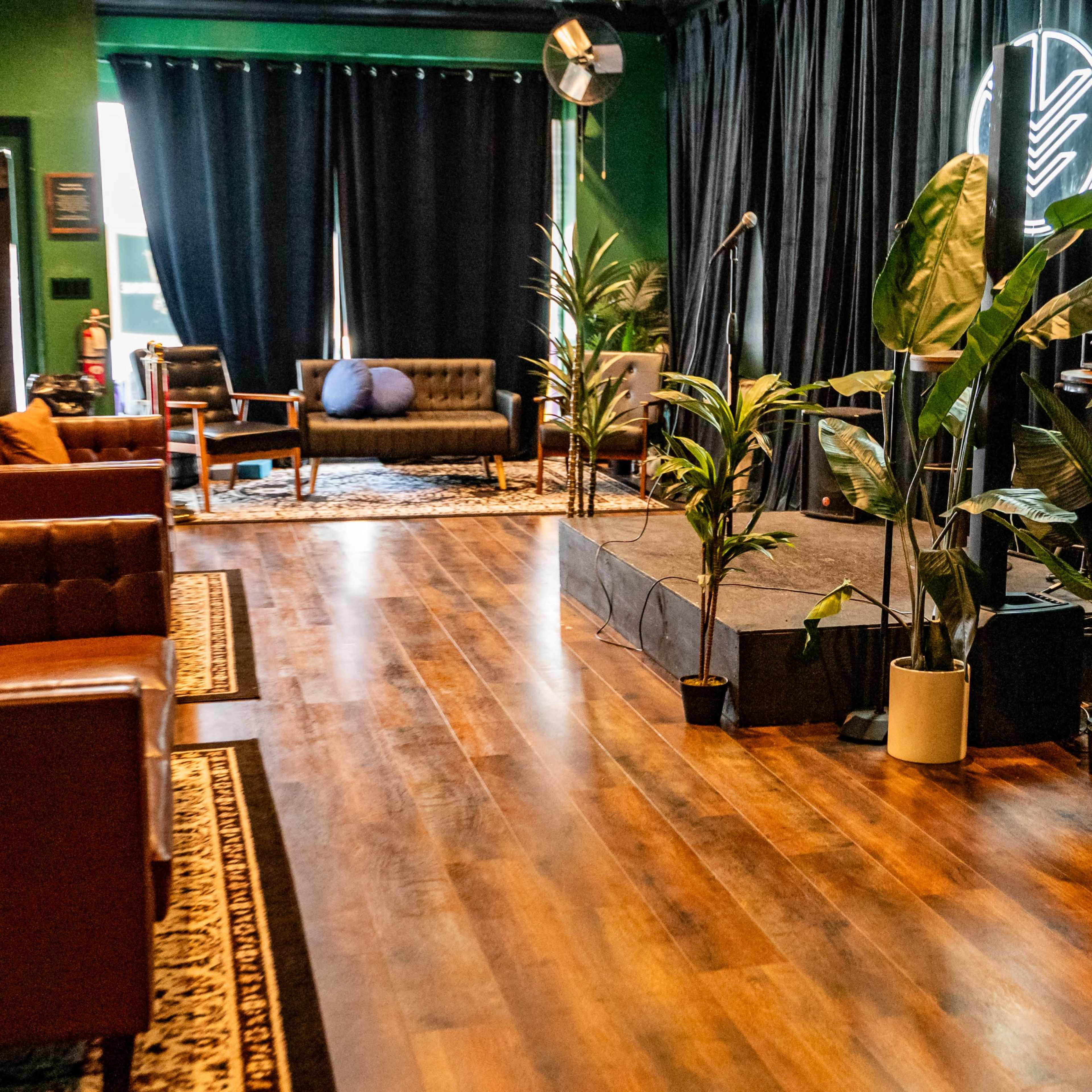 A cozy performance space with brown leather sofas, potted plants, and a small stage illuminated against a dark backdrop.