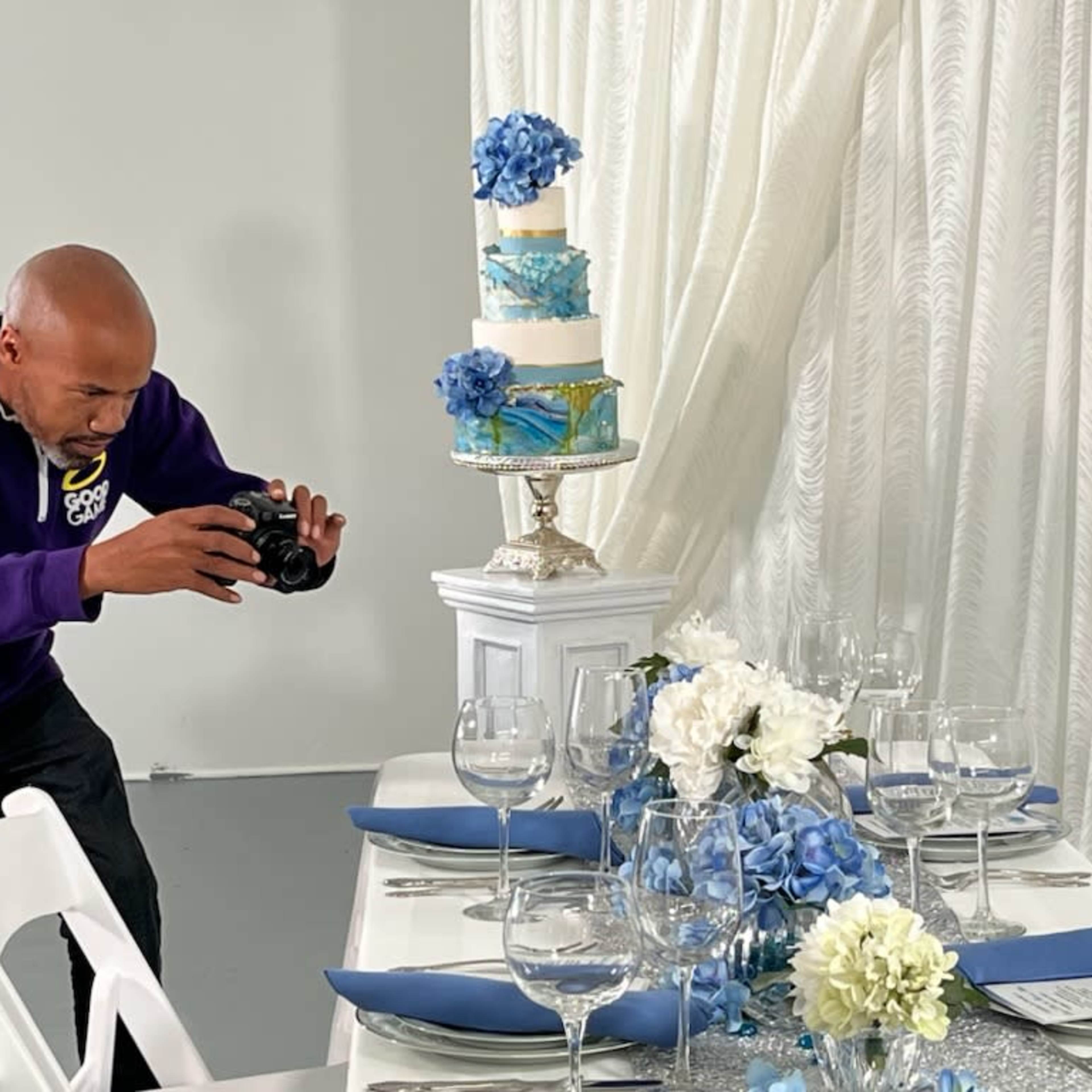 A man in a purple hoodie is taking a photo of a decorated cake on a pedestal at a table set with blue napkins and floral arrangements.