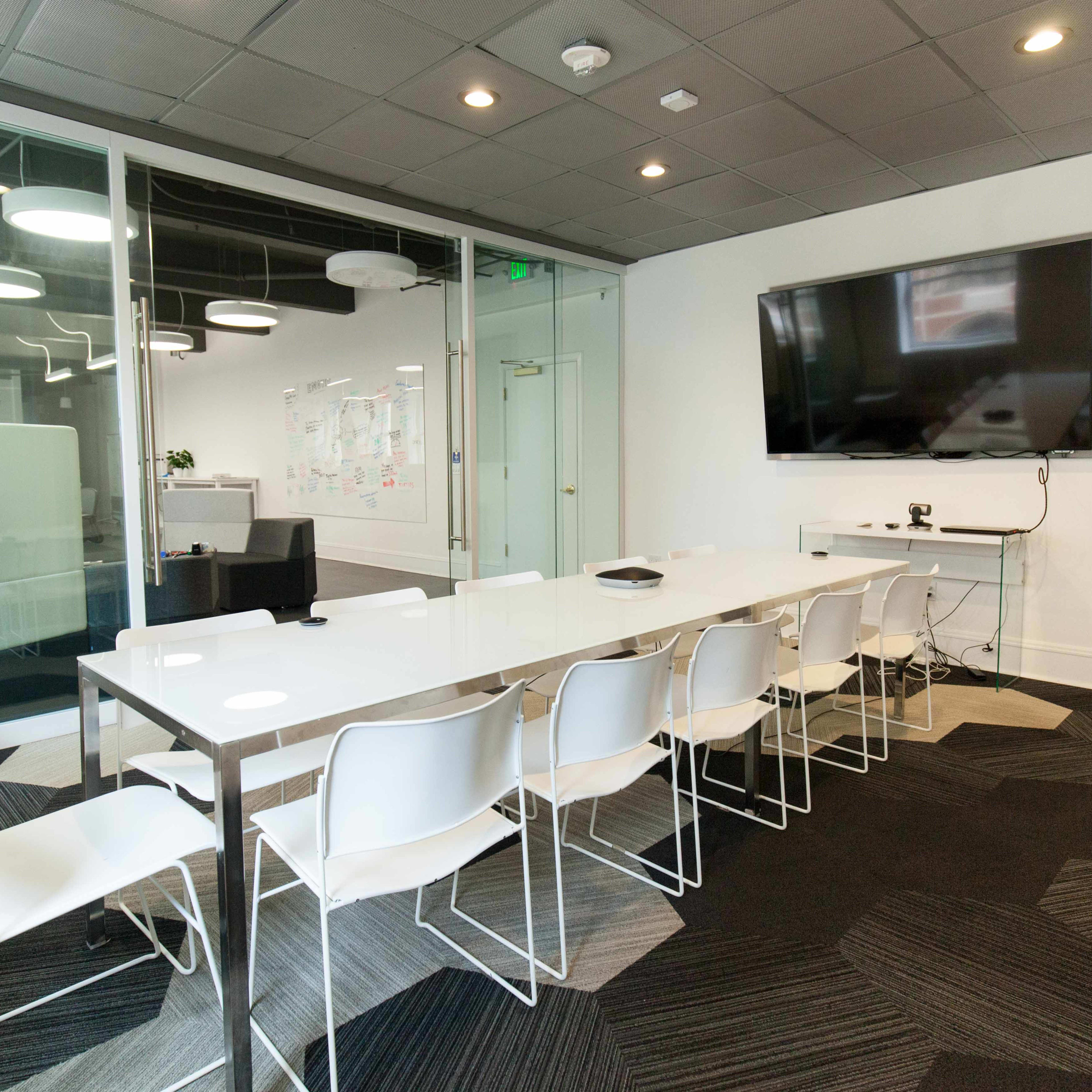 A modern conference room features a long white table with ten white chairs, a large wall-mounted screen, and glass walls displaying a collaborative space.