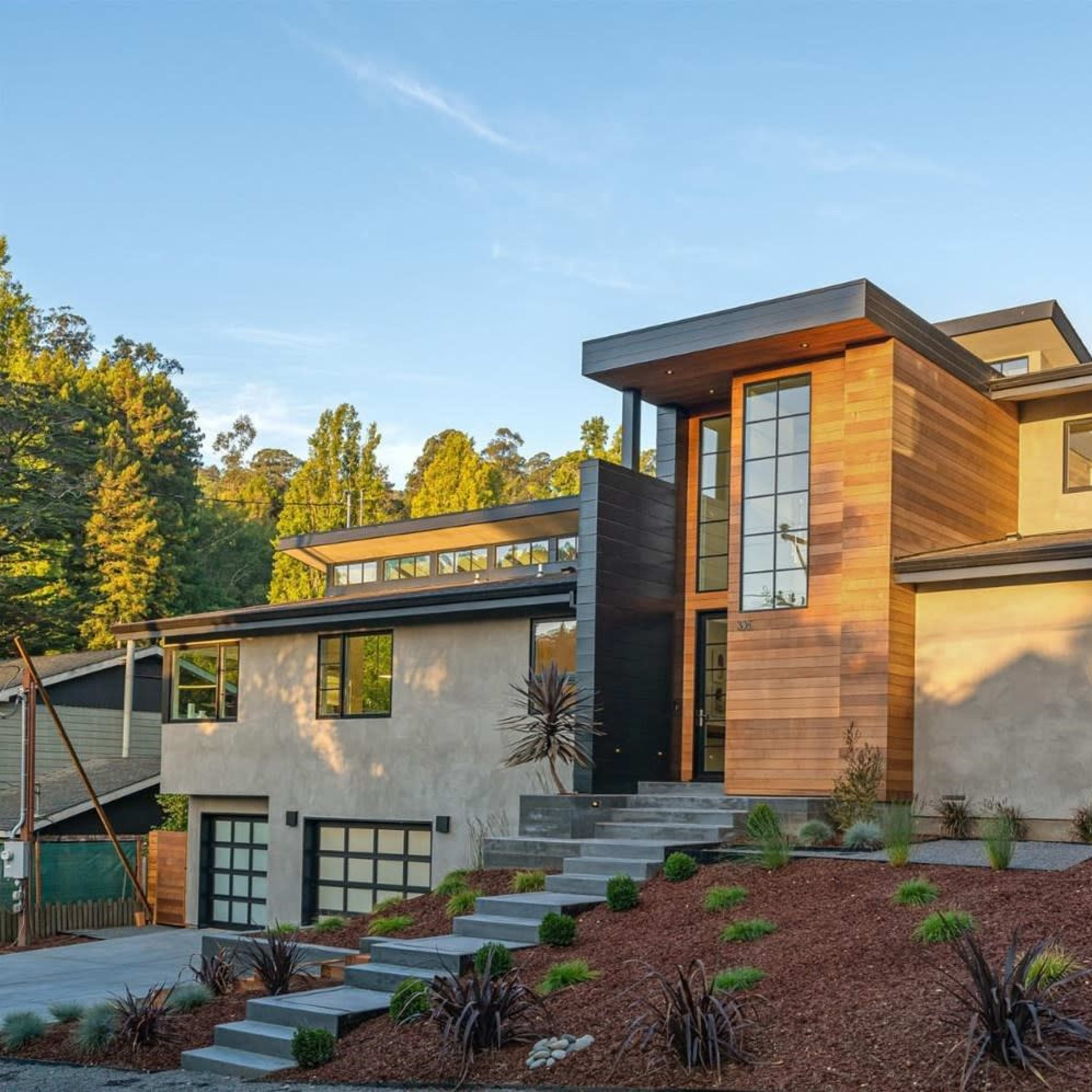 A modern two-story house with a wooden and concrete facade, surrounded by landscaped greenery and a stone pathway.