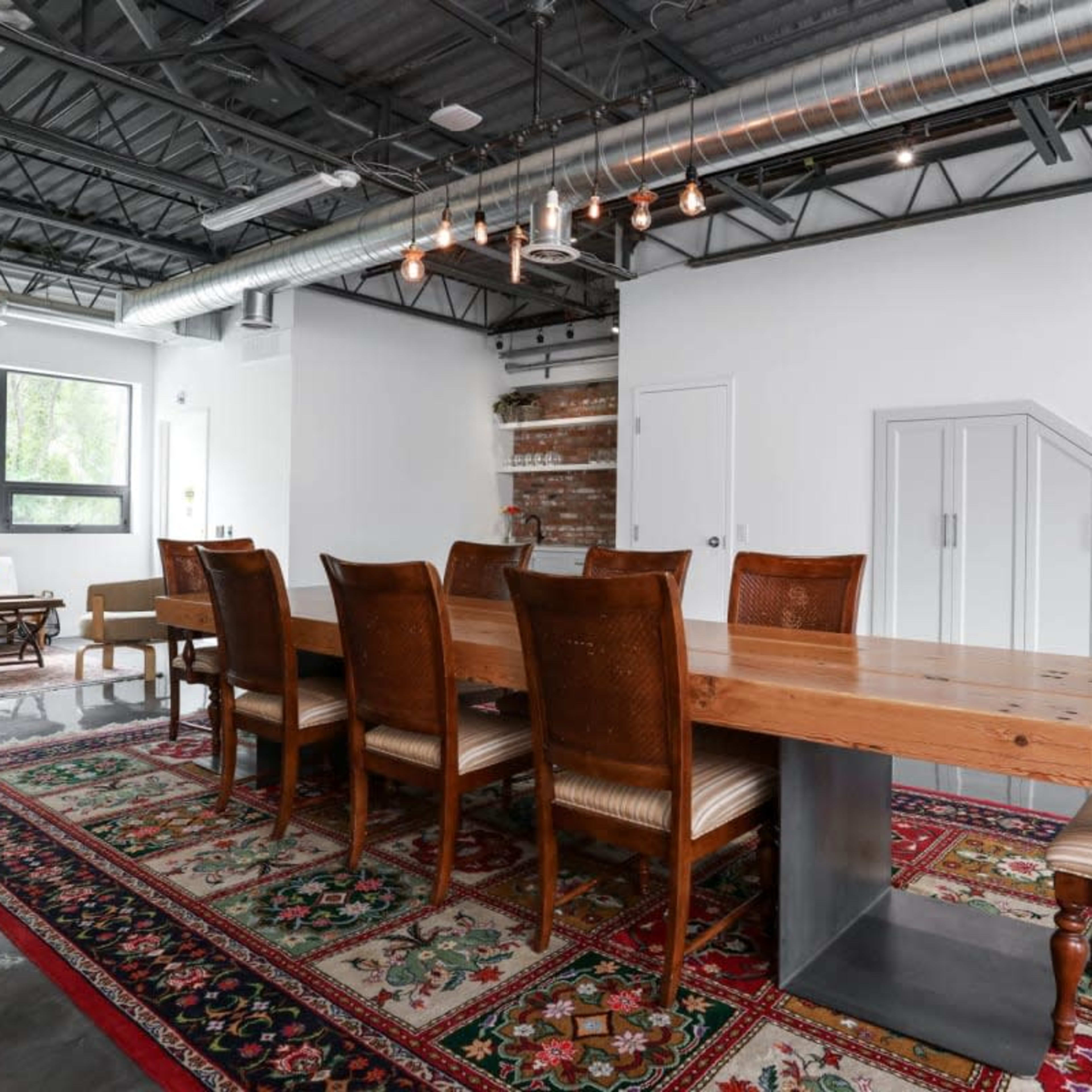 A long wooden dining table with eight chairs is centered on a decorative rug in a modern, industrial-style room featuring exposed metal beams and large windows.