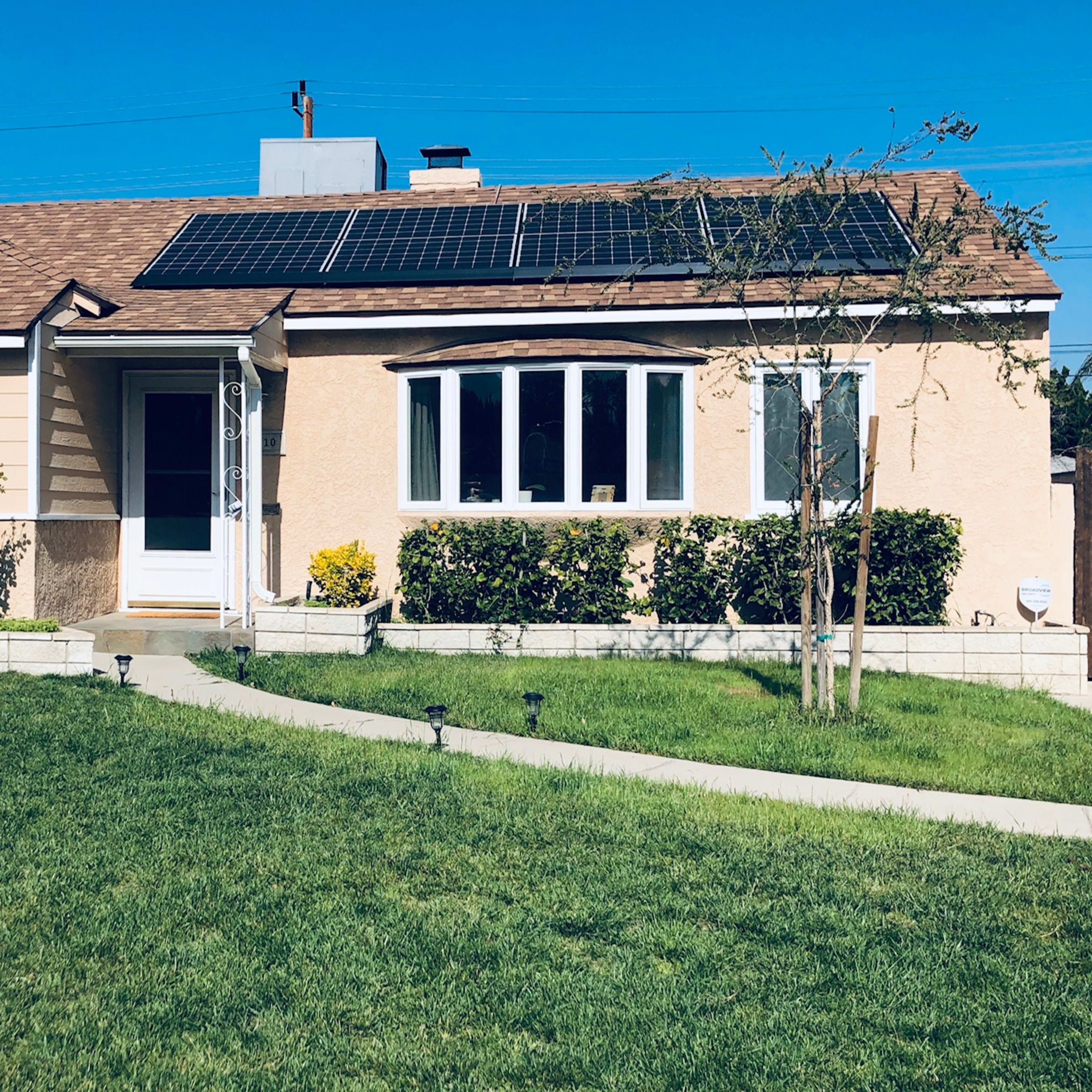 A single-story house with solar panels on the roof is surrounded by a green lawn and flower beds.
