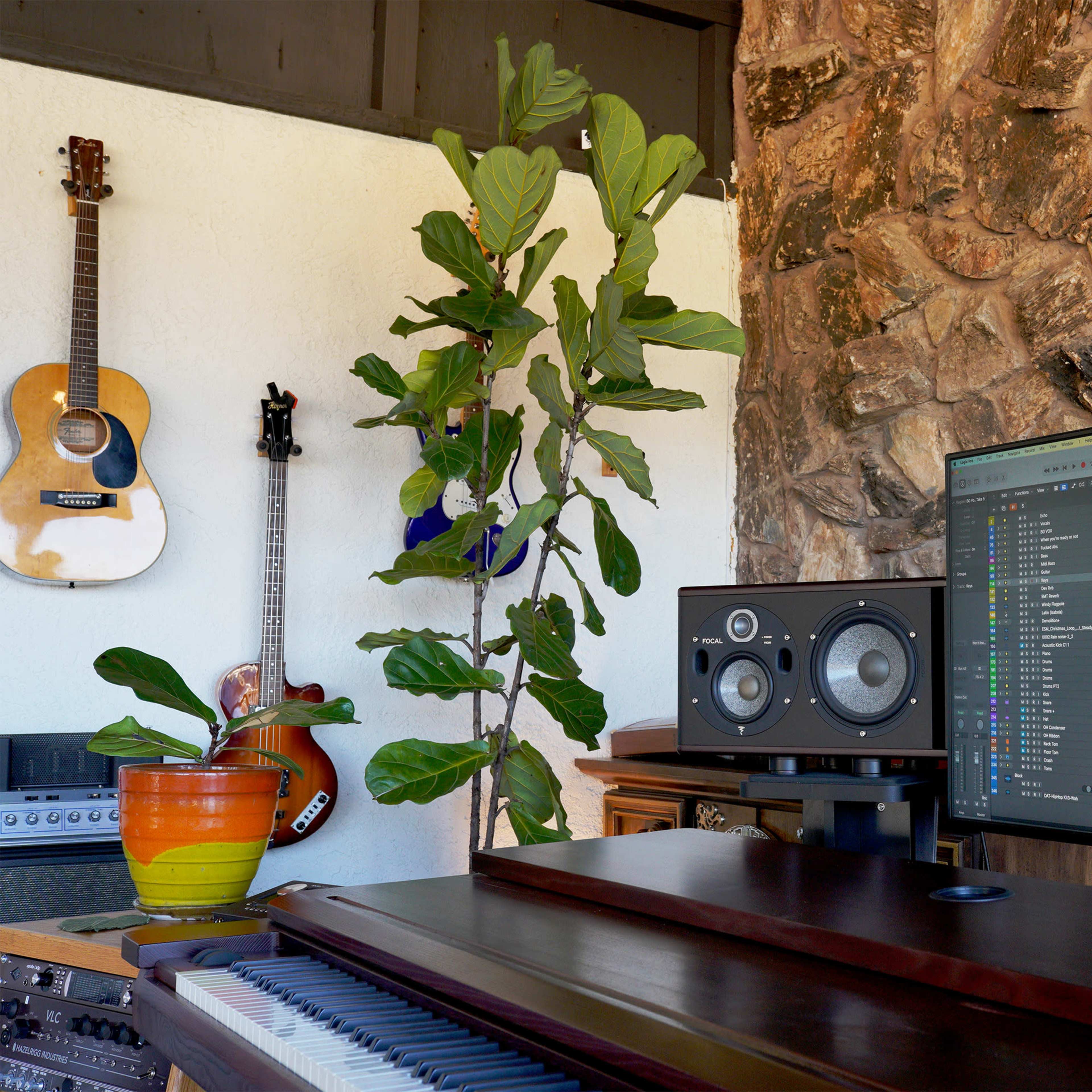 A music production setup features a large monitor displaying audio software, surrounded by guitars, a potted plant, and studio speakers.