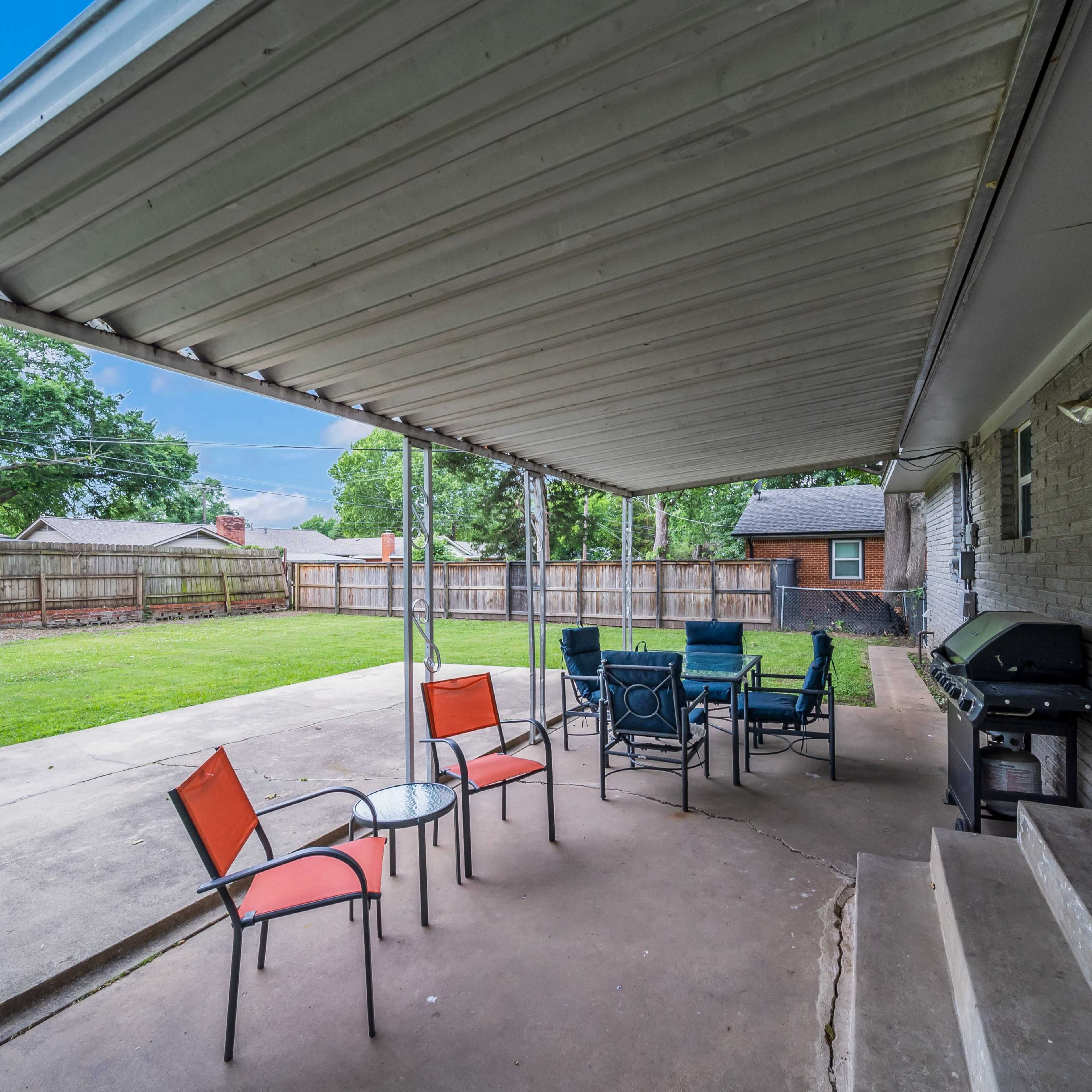 A covered patio area with several chairs and a table, overlooking a grassy backyard enclosed by a fence.