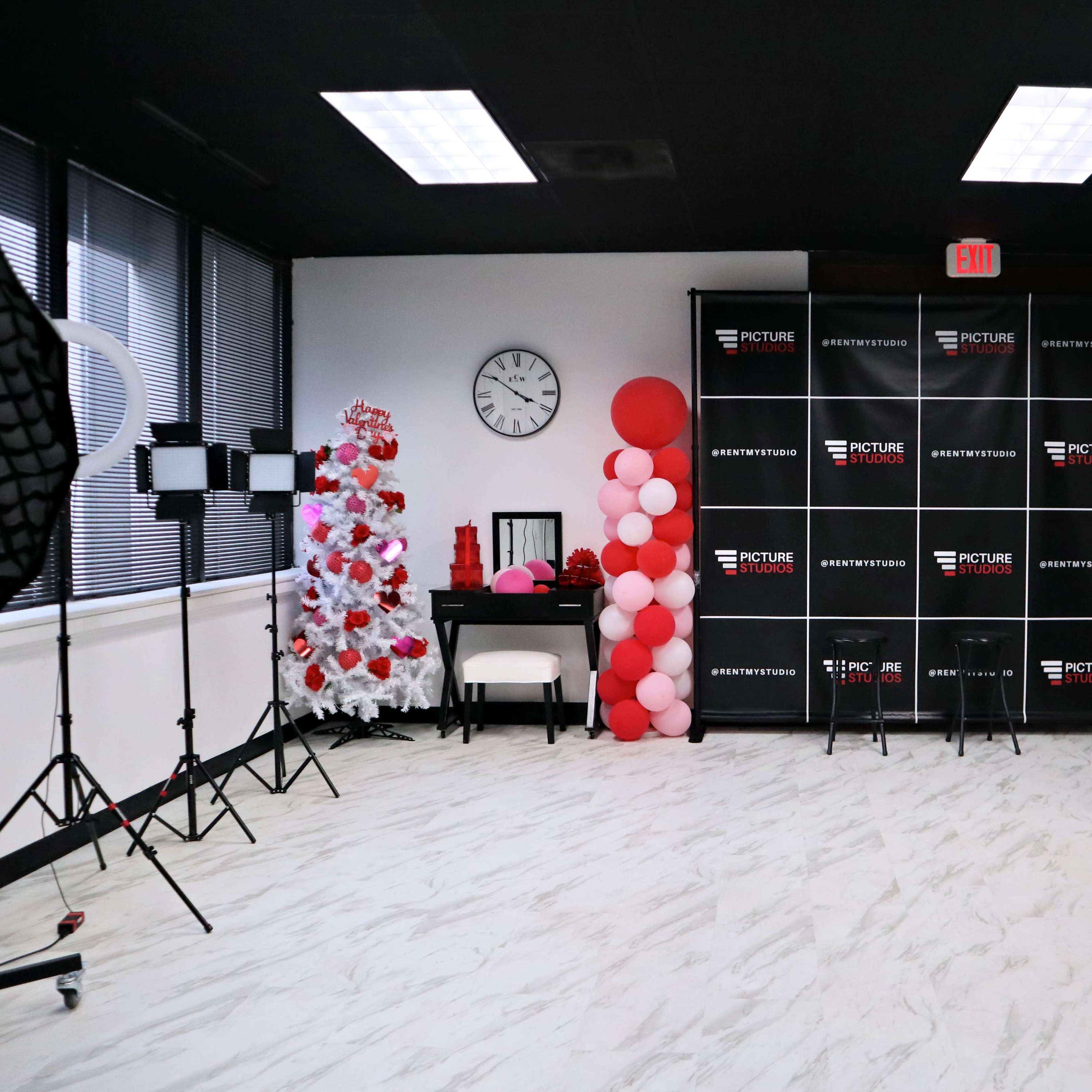 A photo studio setup featuring a decorative backdrop with black and white panels, a clock on the wall, lighting equipment, and a display of balloons in red and pink hues.