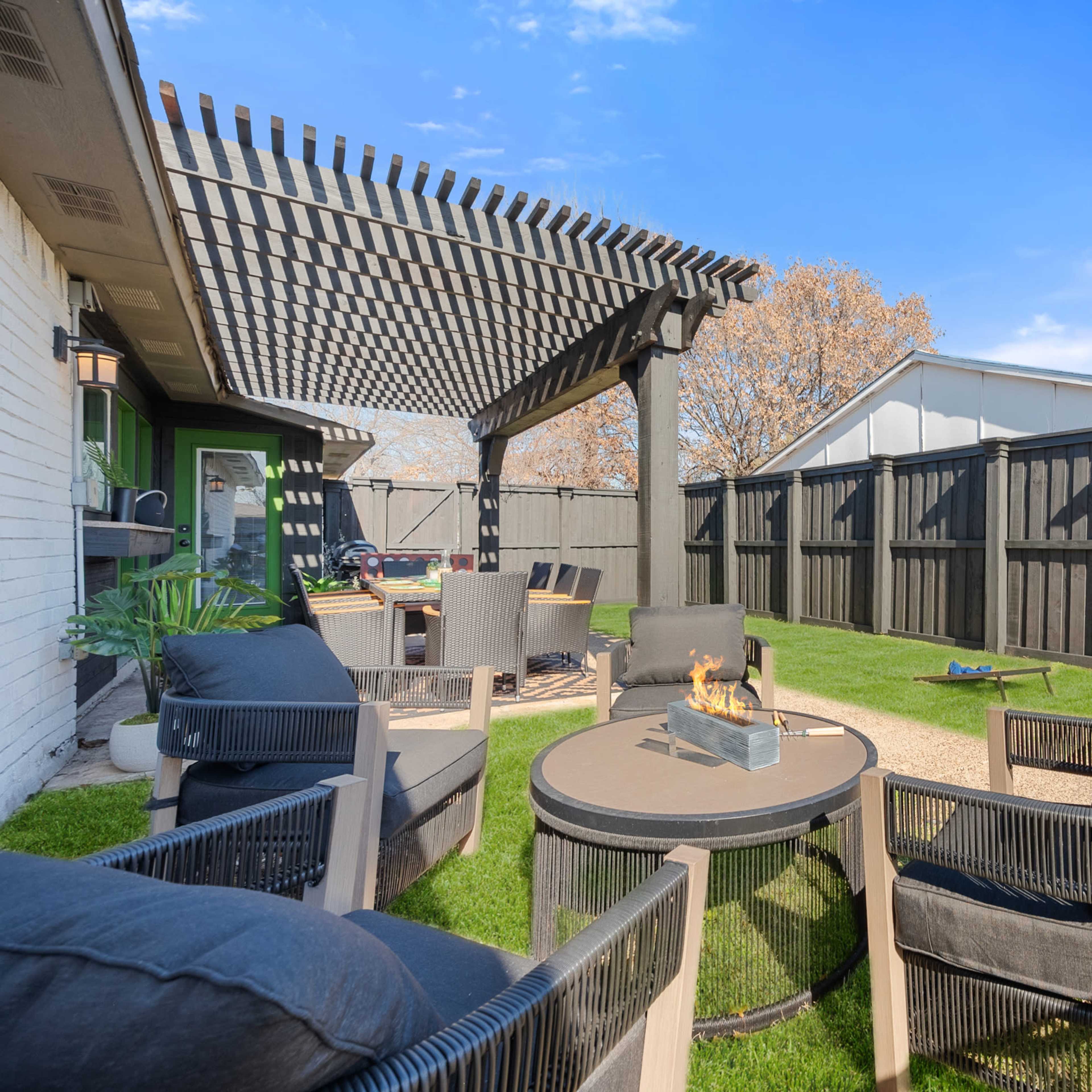 A backyard seating area features a fire pit surrounded by comfortable chairs under a wooden pergola.