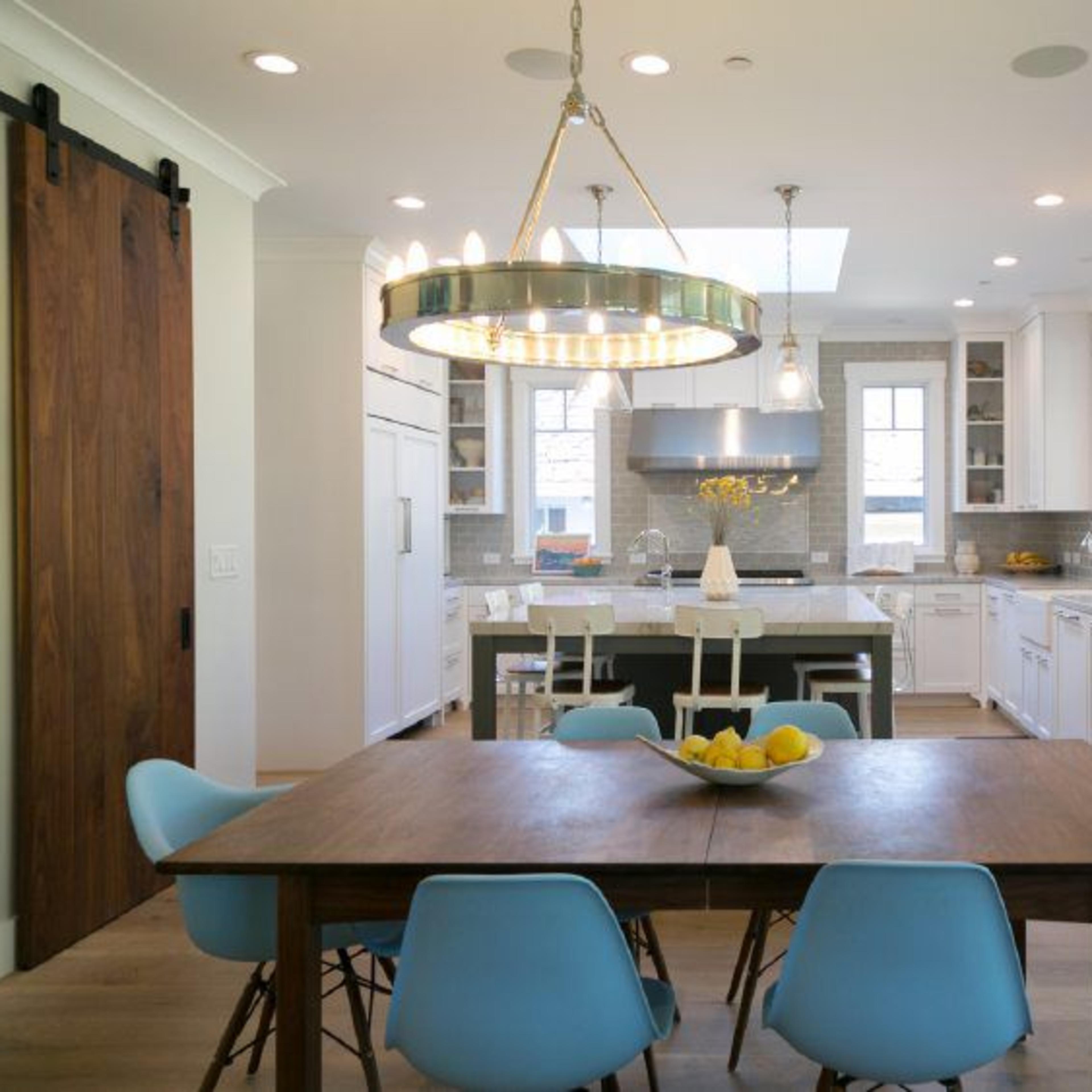 A modern kitchen features a large wooden dining table surrounded by blue chairs, with a chandelier overhead and white cabinetry in the background.