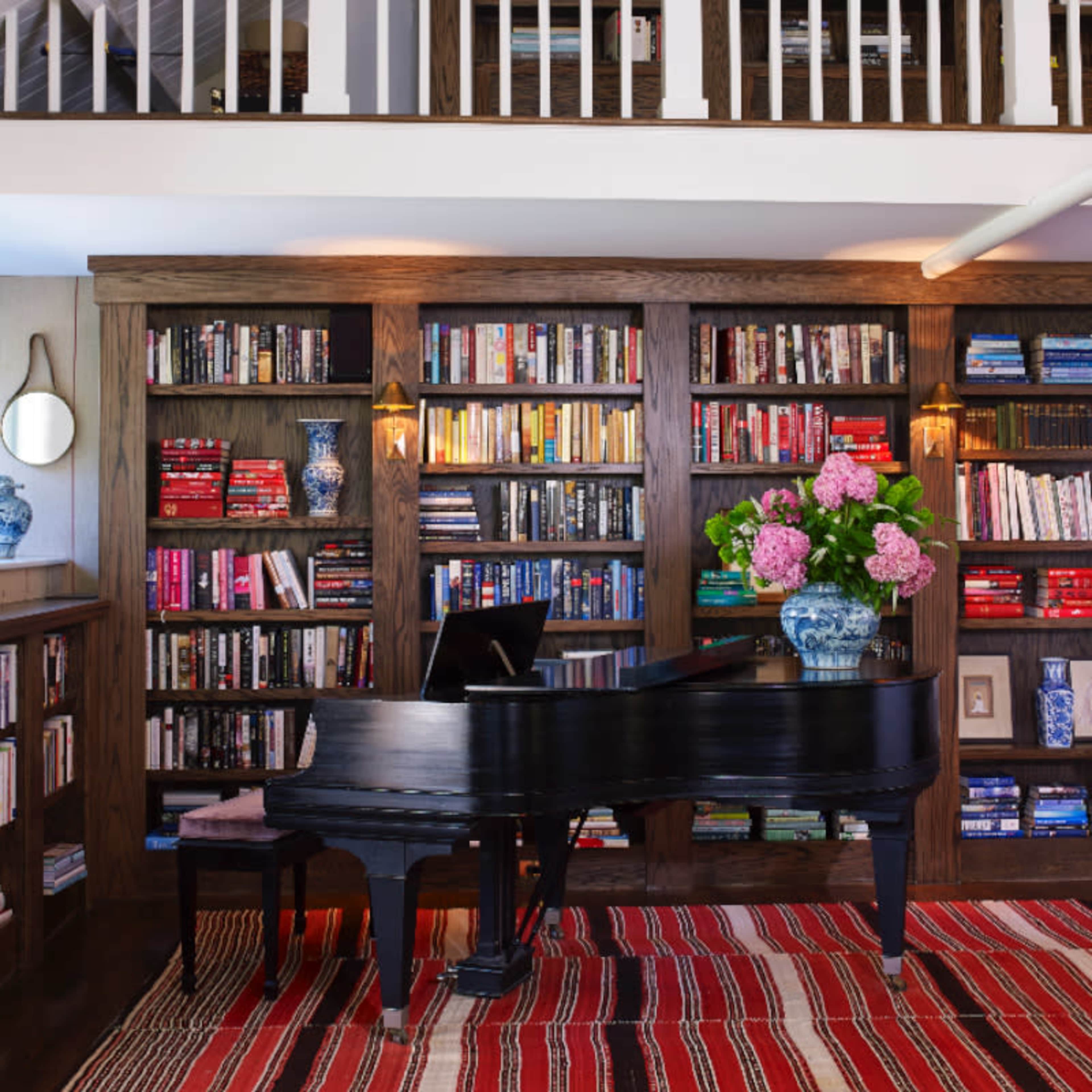 A black grand piano sits beside a floral arrangement in a room featuring a wooden bookshelf filled with colorful books and decorative items.