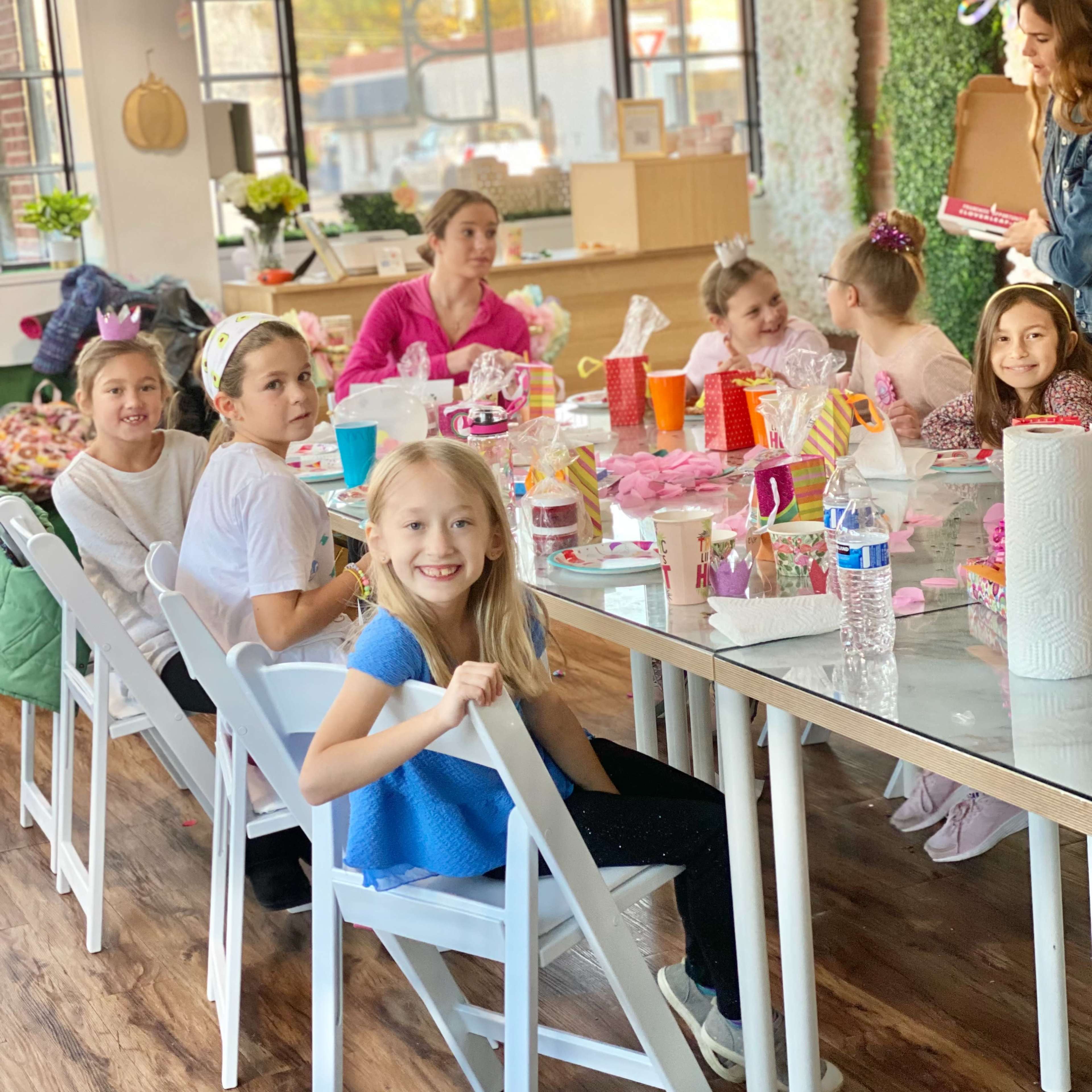 A group of children and adults are seated at a table covered with party supplies in a brightly lit event space.