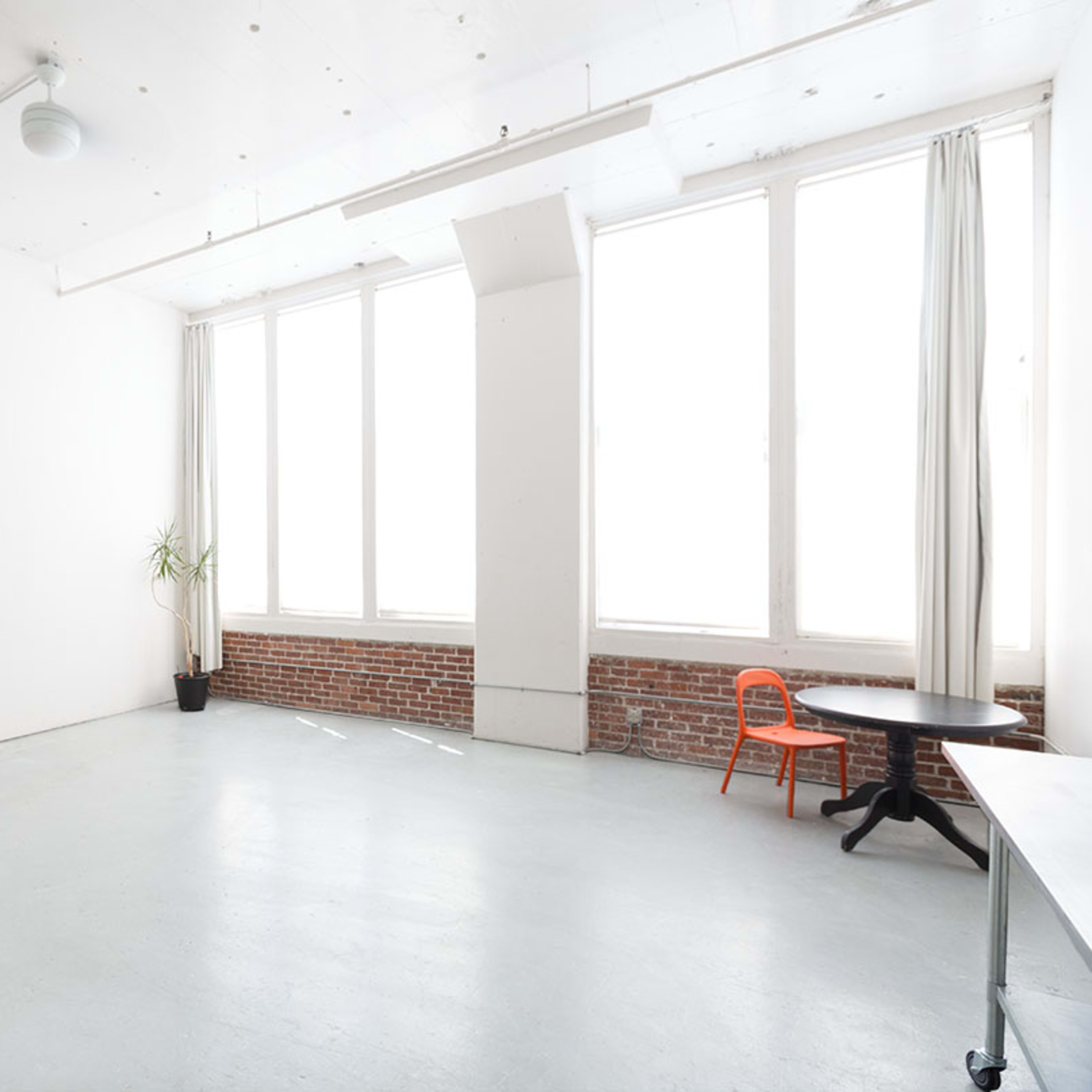 A spacious, empty room with large windows, a brick wall, and minimal furniture including an orange chair and a table.