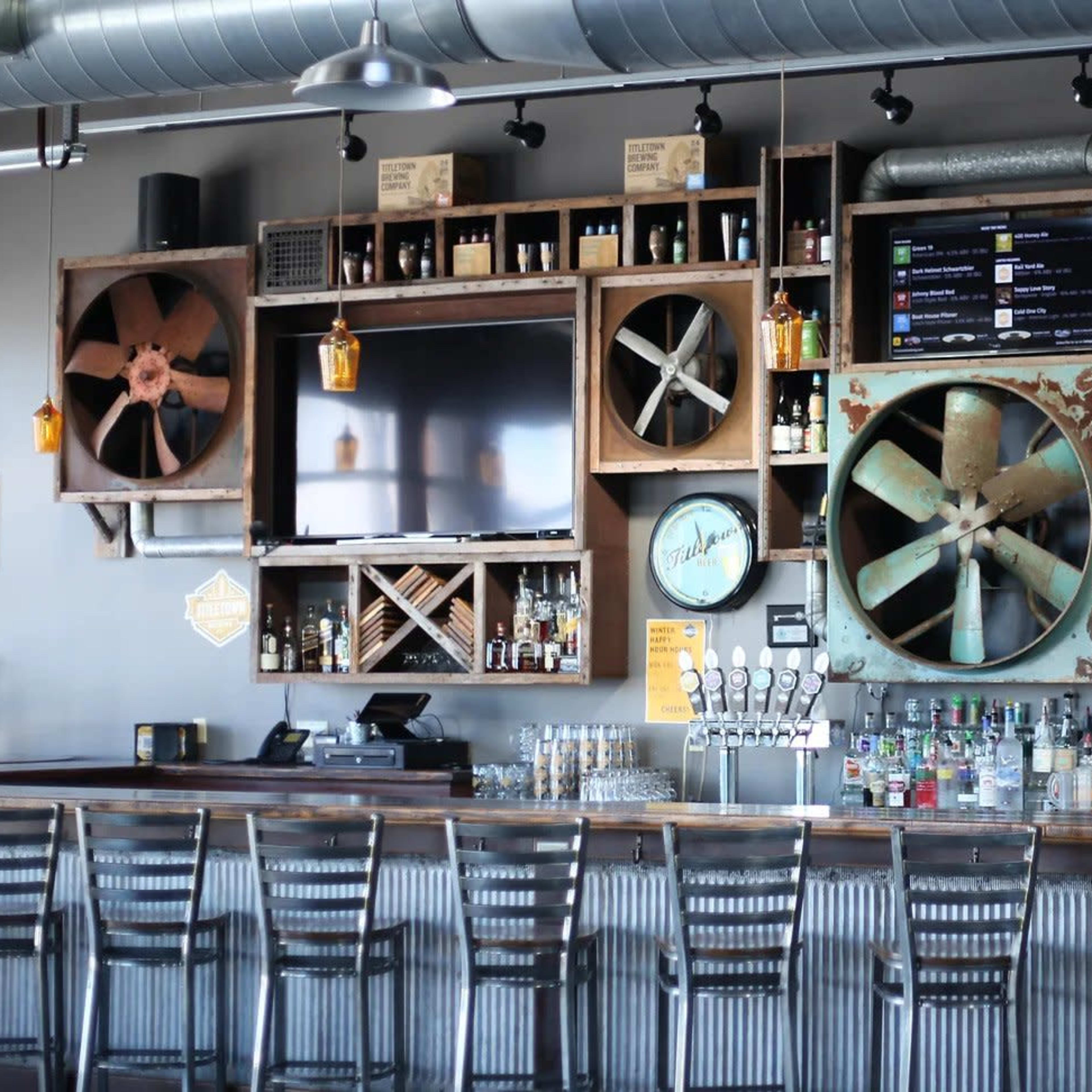 A trendy bar interior with a rustic wooden wall decorated with large fan replicas, televisions, and various bottles of liquor arranged on shelves.
