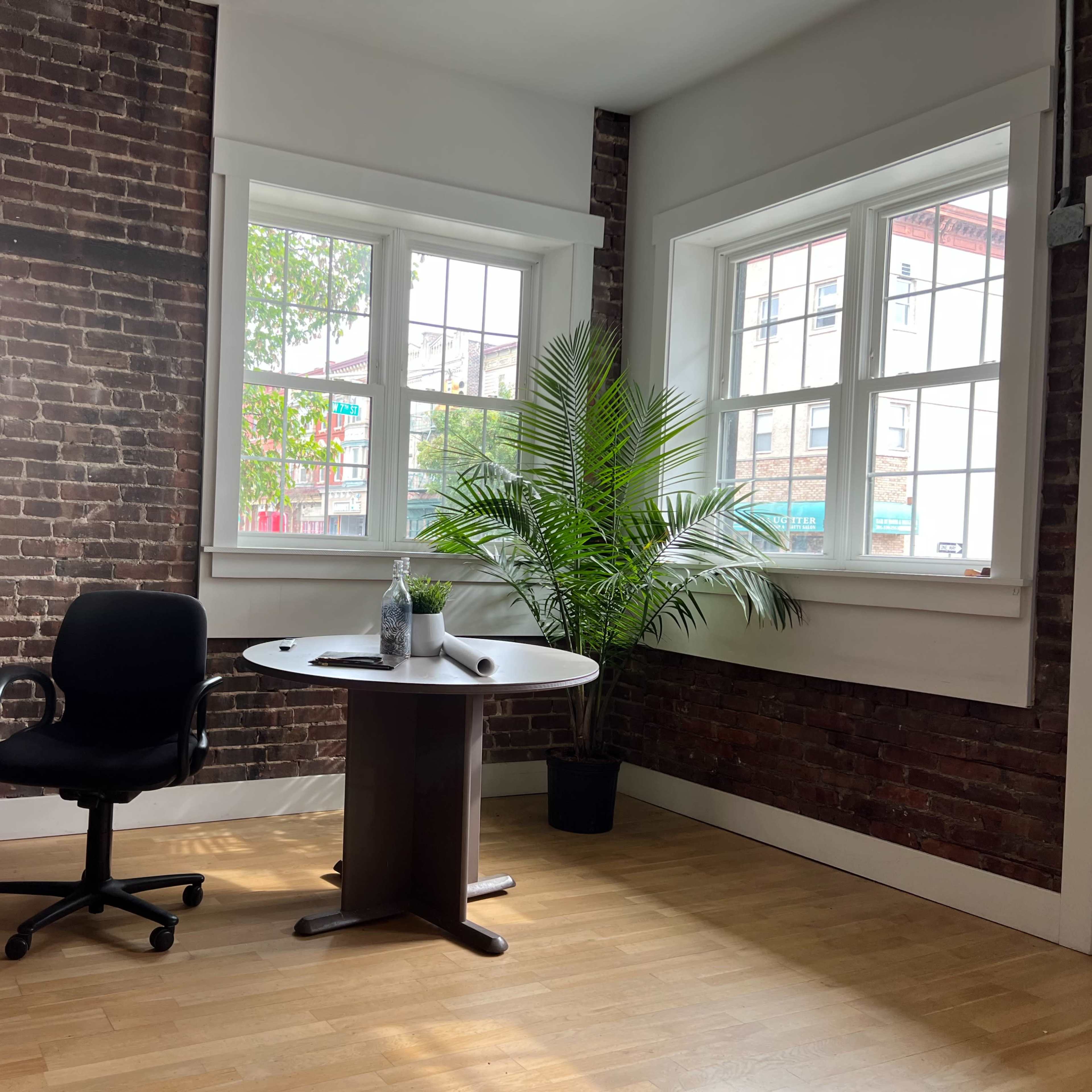 An empty office space with a black swivel chair and a round table positioned near two large windows, against a backdrop of exposed brick walls and a potted plant.