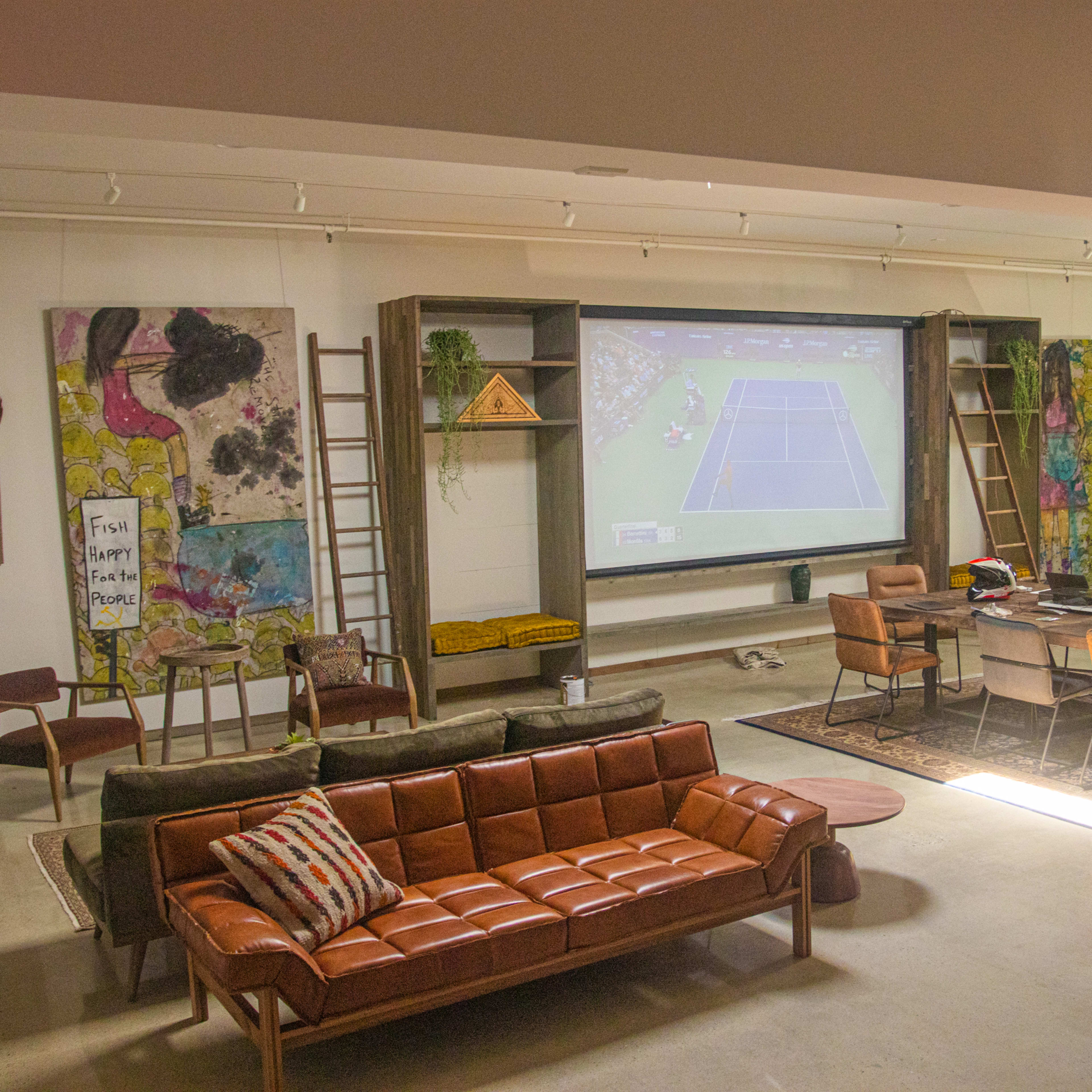 A spacious room with a large screen displaying a tennis match, surrounded by modern furniture, artwork on the walls, and a dining table set for a meal.