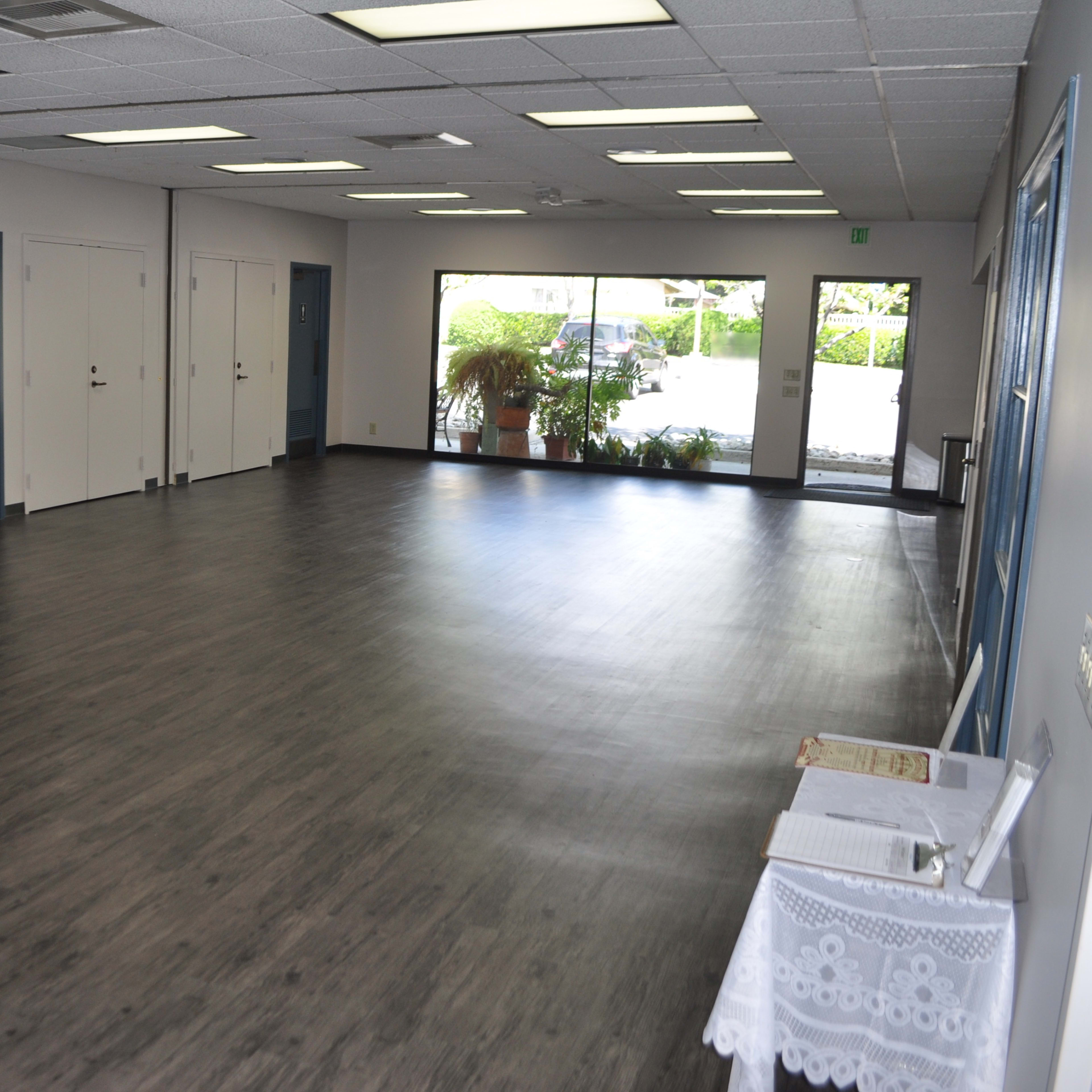 A large, empty indoor space with light-colored walls and a wooden floor, featuring several closed doors and a window that leads to an outdoor area.