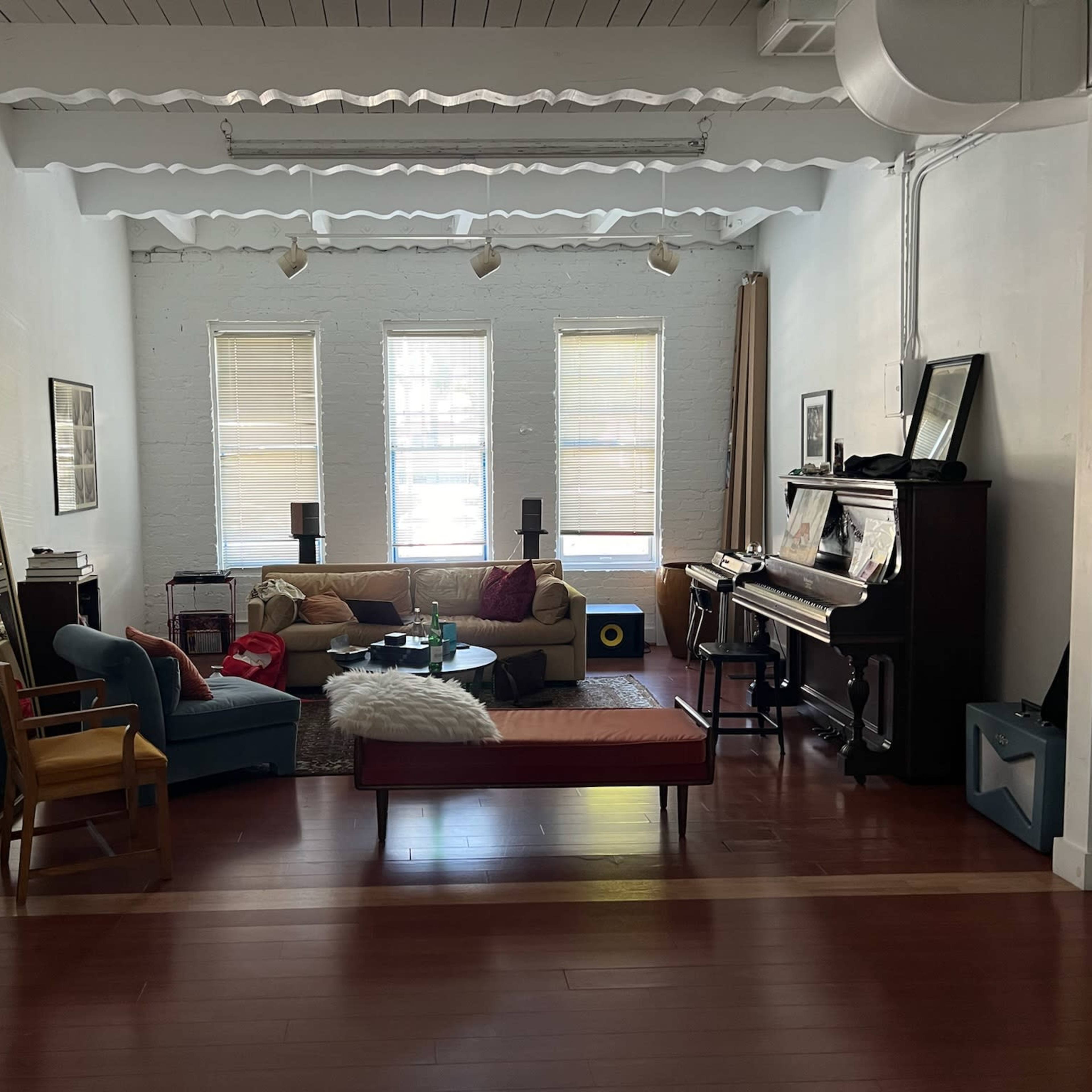 A spacious living room with a sofa, chair, and a piano, featuring large windows and wooden floors.