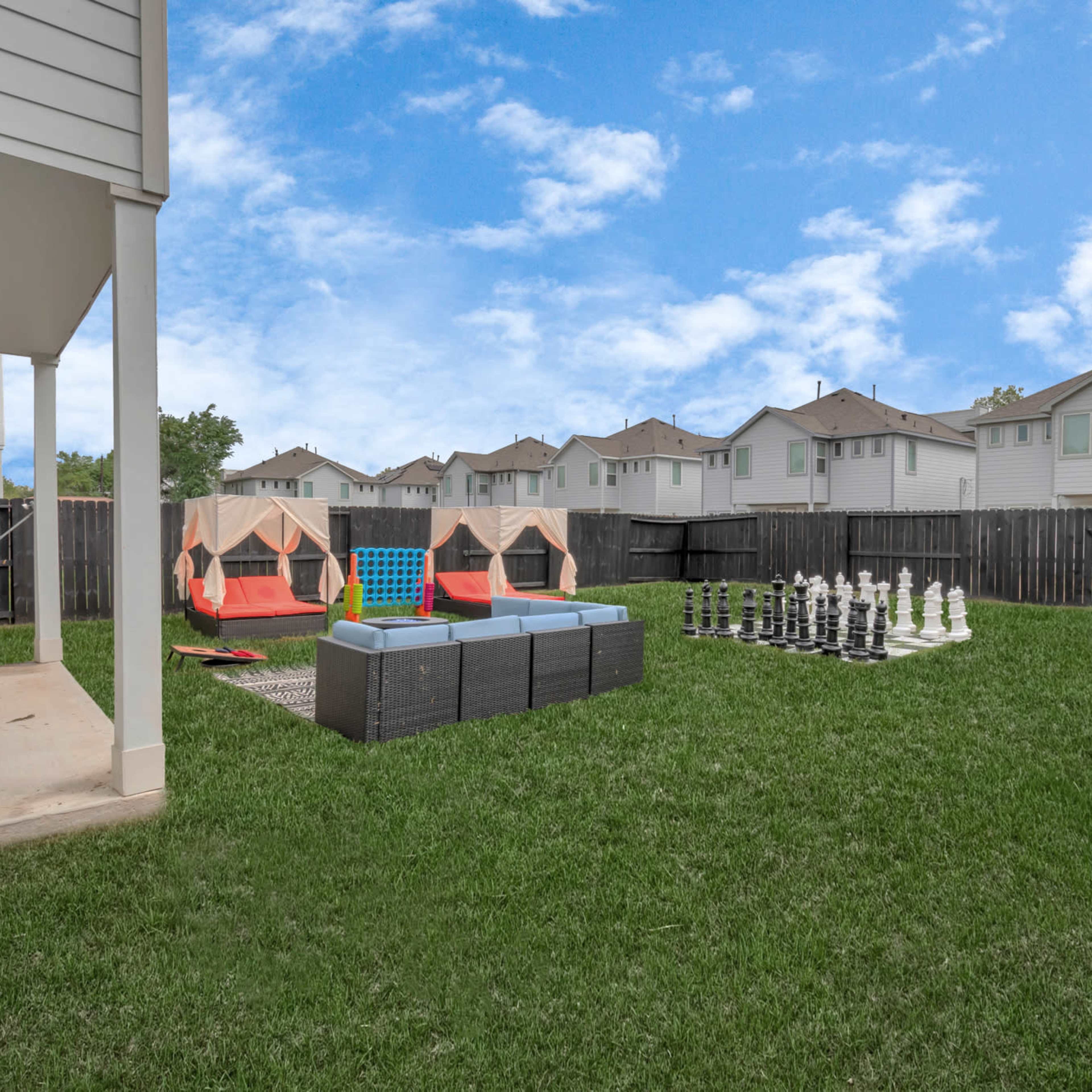 A backyard with two red and blue lounge chairs, a large chess set, and several tents, surrounded by a green lawn and lined by wooden fences.