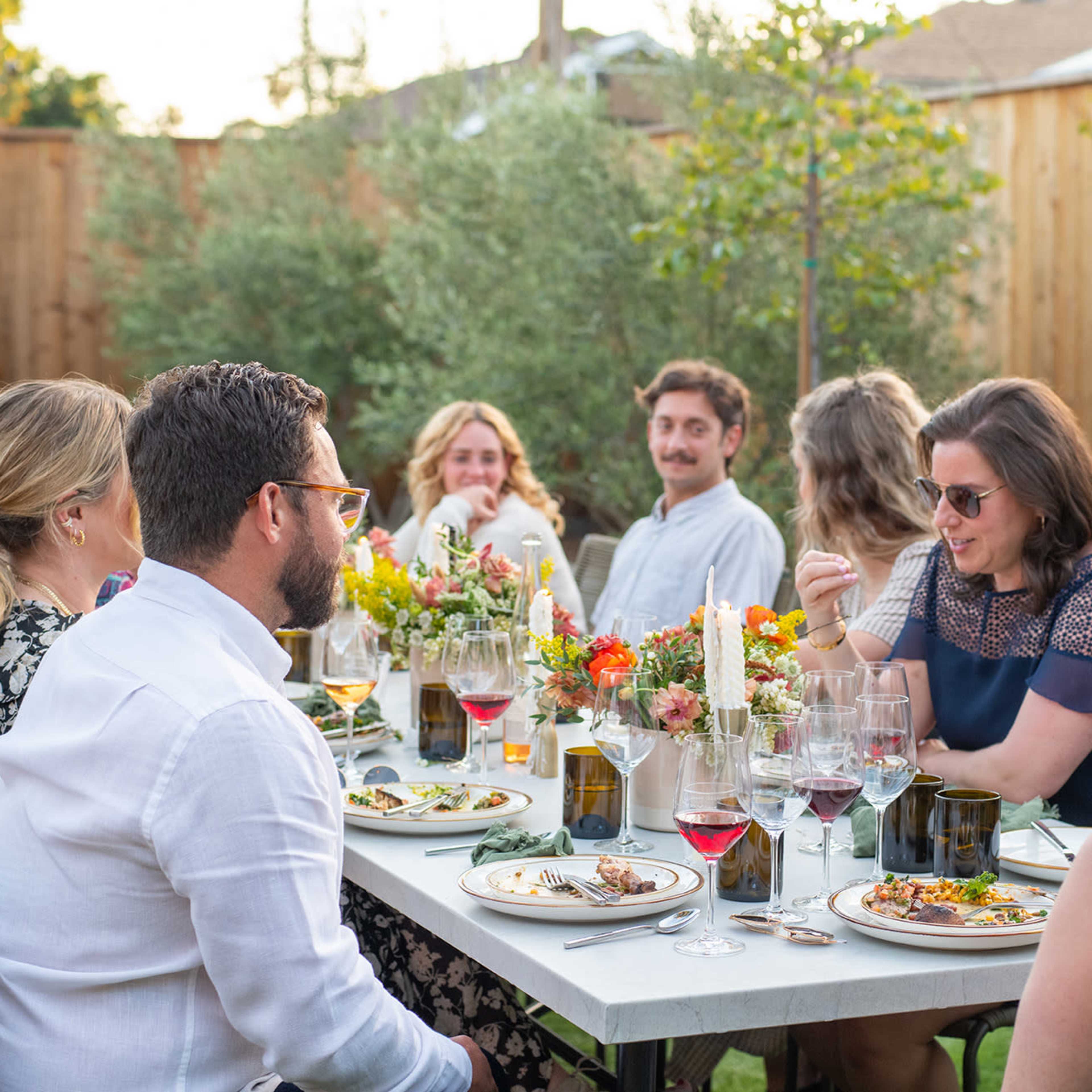 A group of people enjoys a dinner gathering outdoors at a table adorned with floral centerpieces and wine glasses.