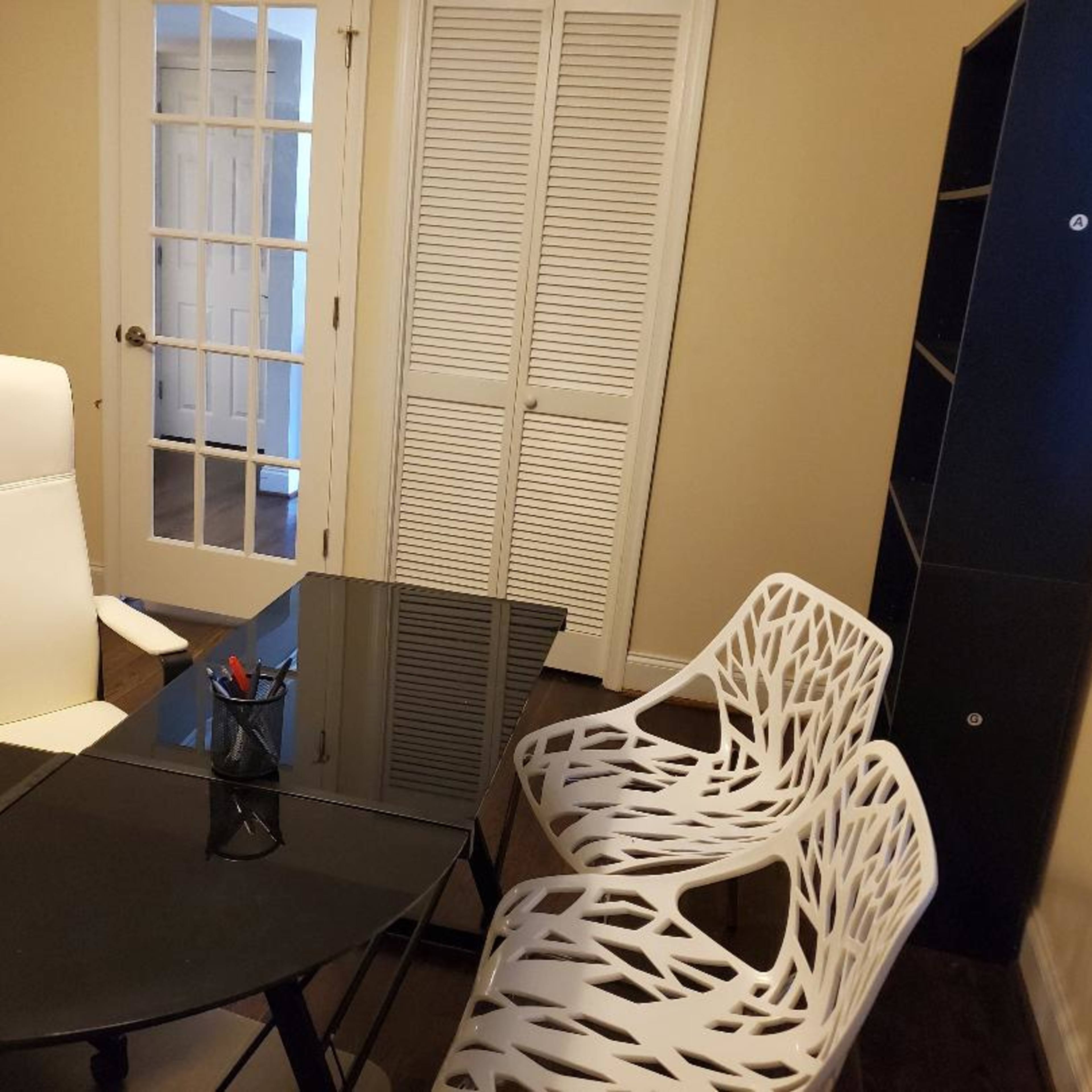 A small office space featuring a black glass table, a white office chair, and two white chairs, with a set of double doors and a storage unit in the background.