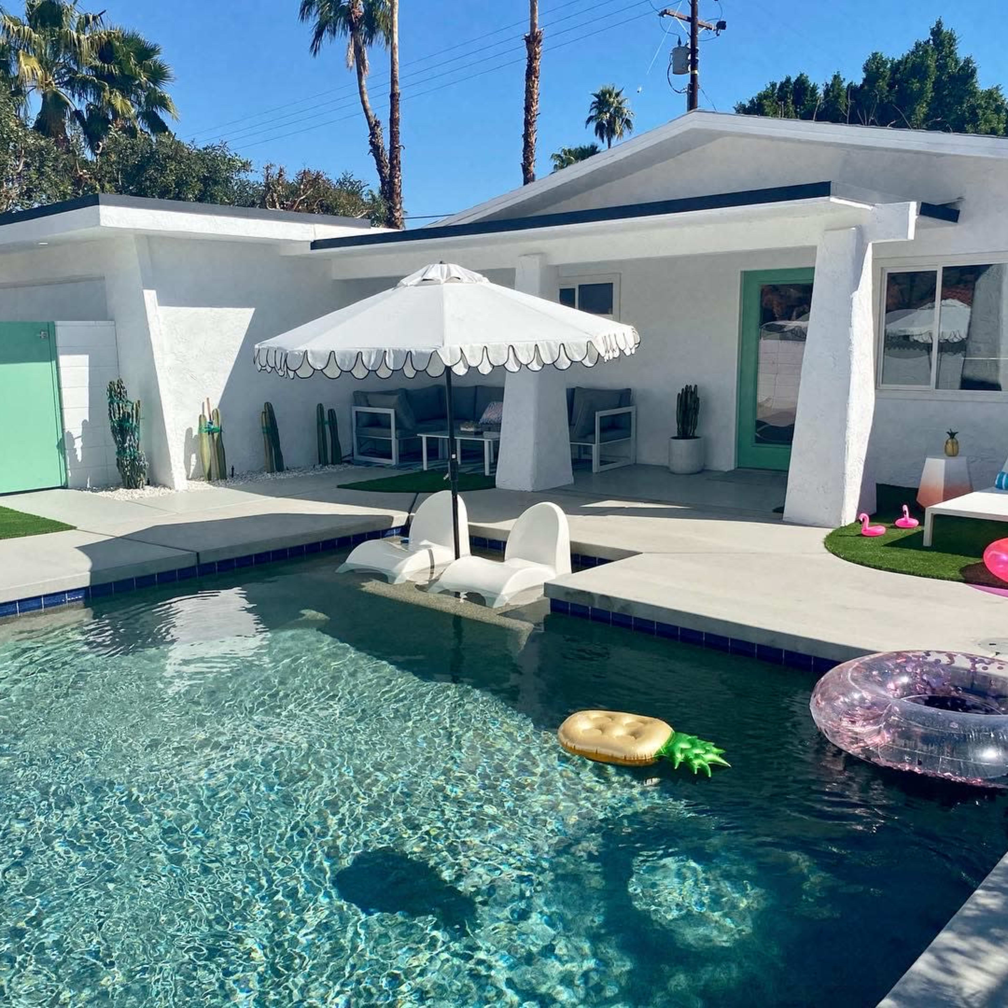A modern backyard features a swimming pool surrounded by lounge chairs, an umbrella, and inflatable toys under clear blue skies.