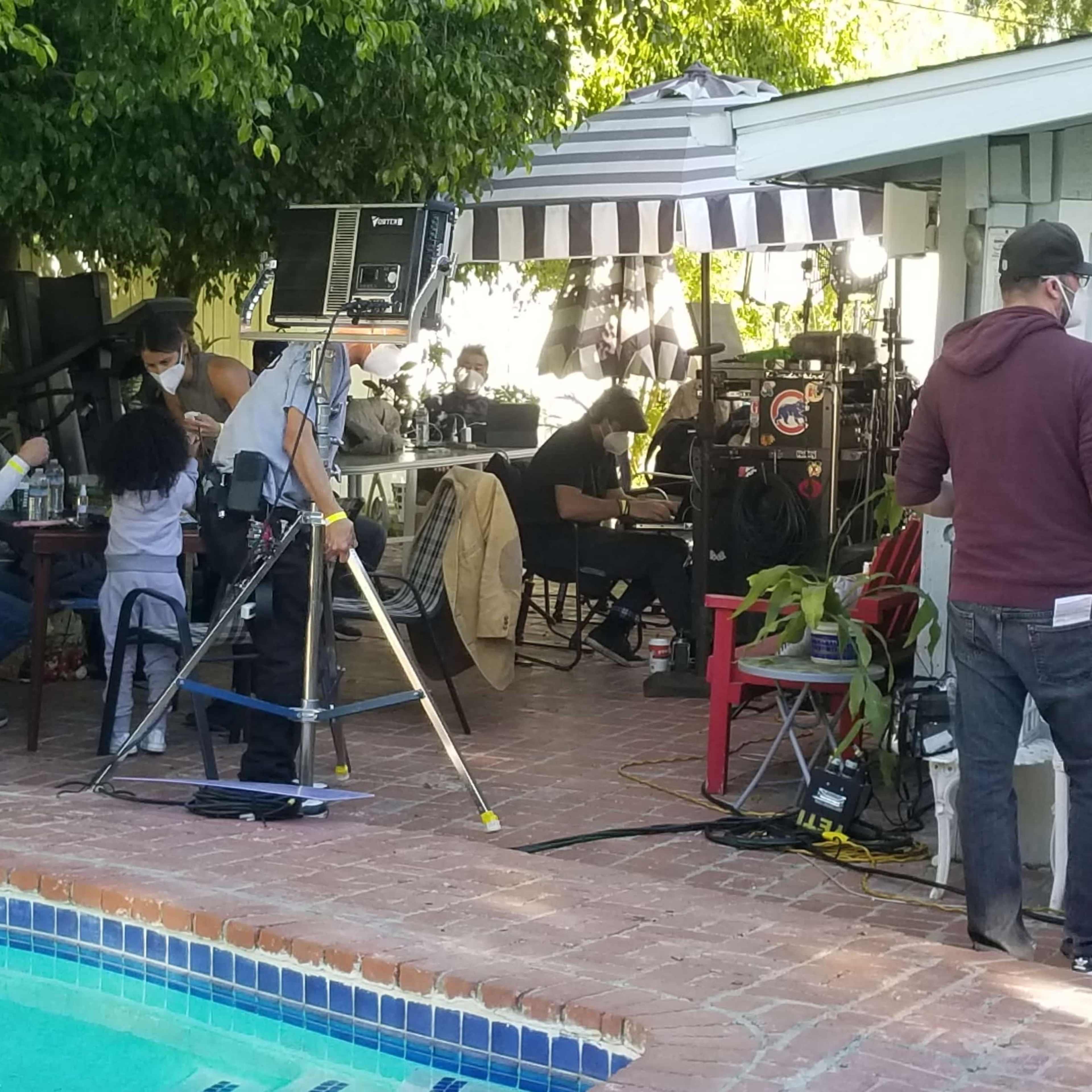 A film crew sets up equipment near a poolside area, with several people gathered around a table and others moving about the scene.