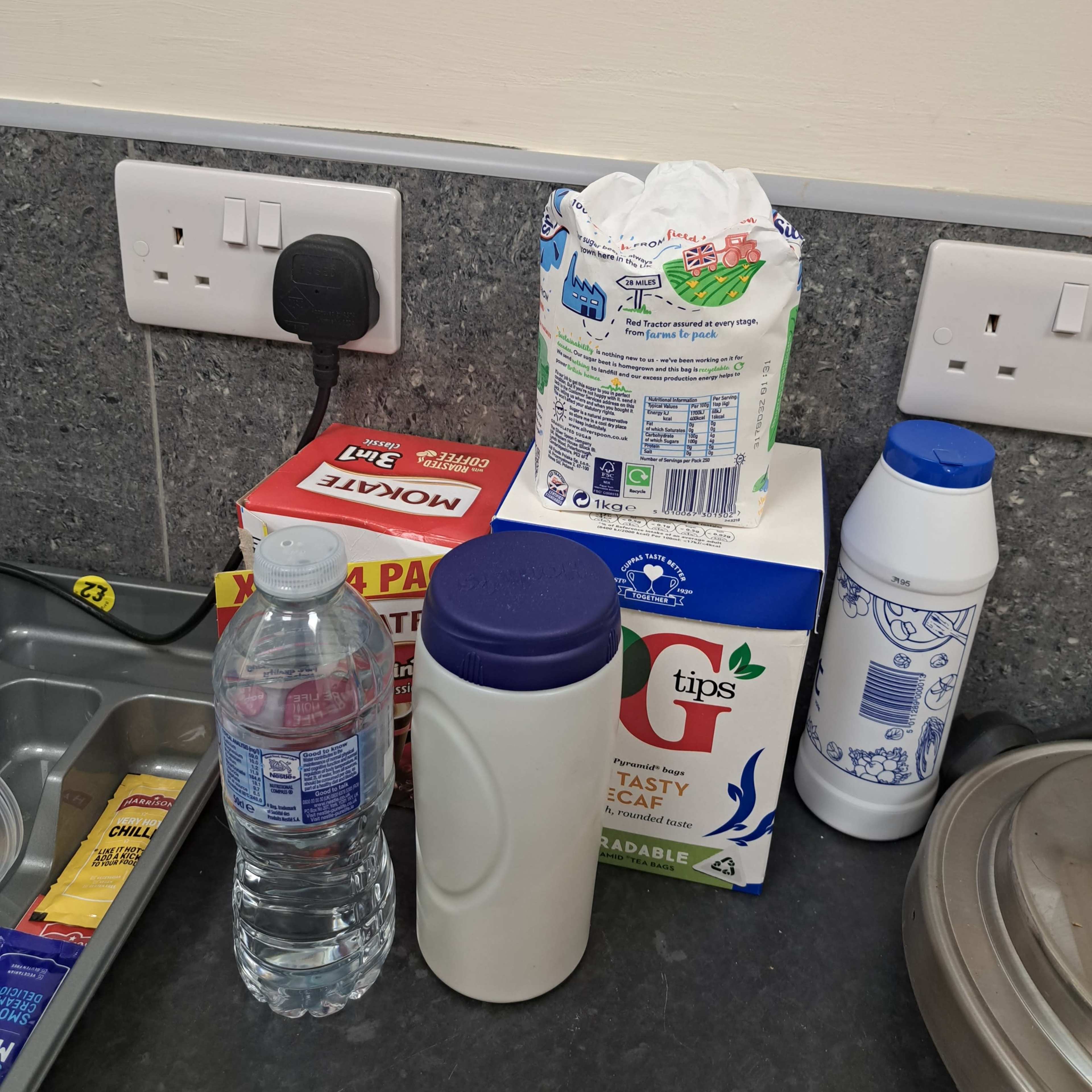 A kitchen counter with a bottle of water, a box of teabags, a container of sugar, a bag of flour, and a plastic container.