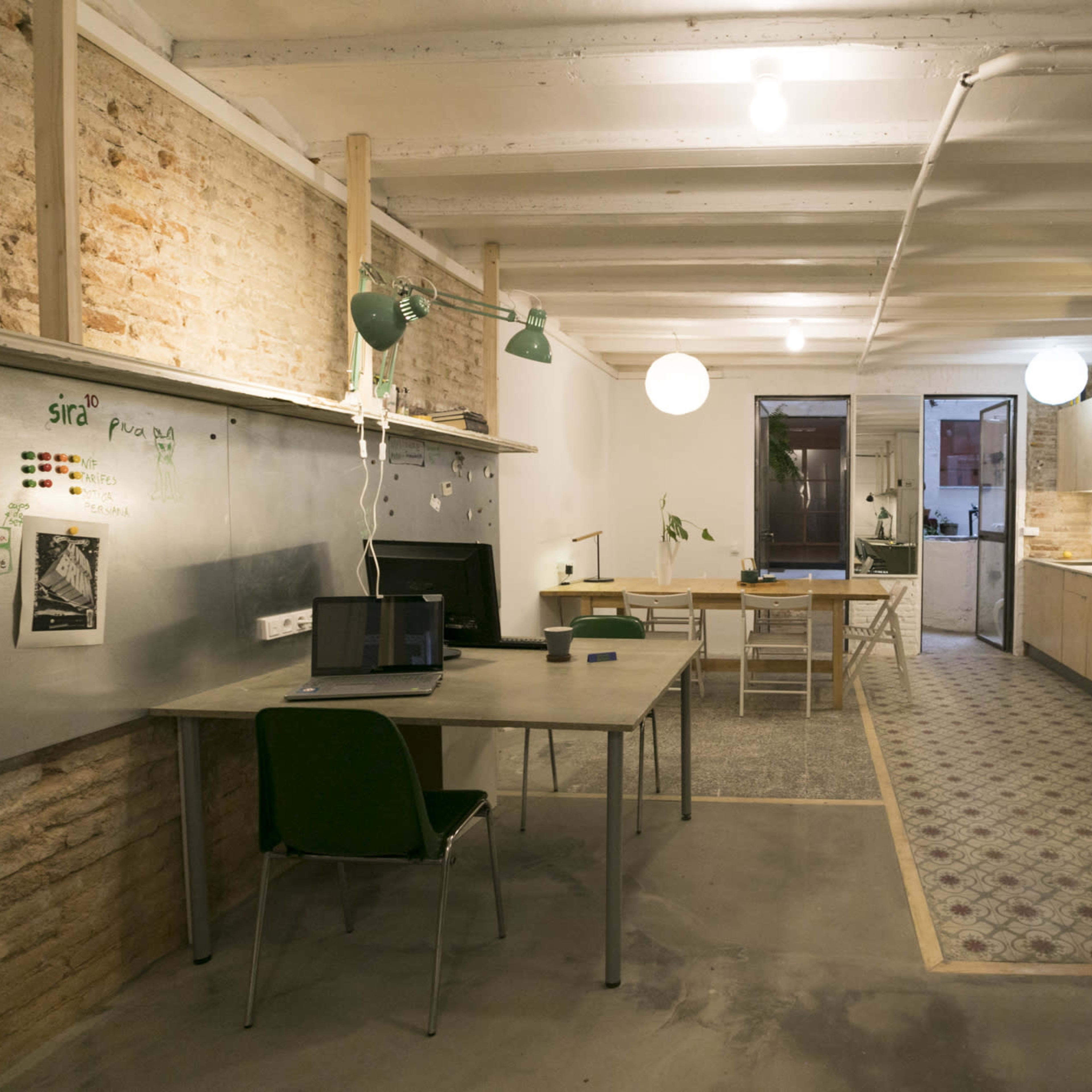 A spacious office with concrete floors, wooden beams, and a mix of desks, laptops, and kitchen areas, illuminated by overhead lights and green lamps.