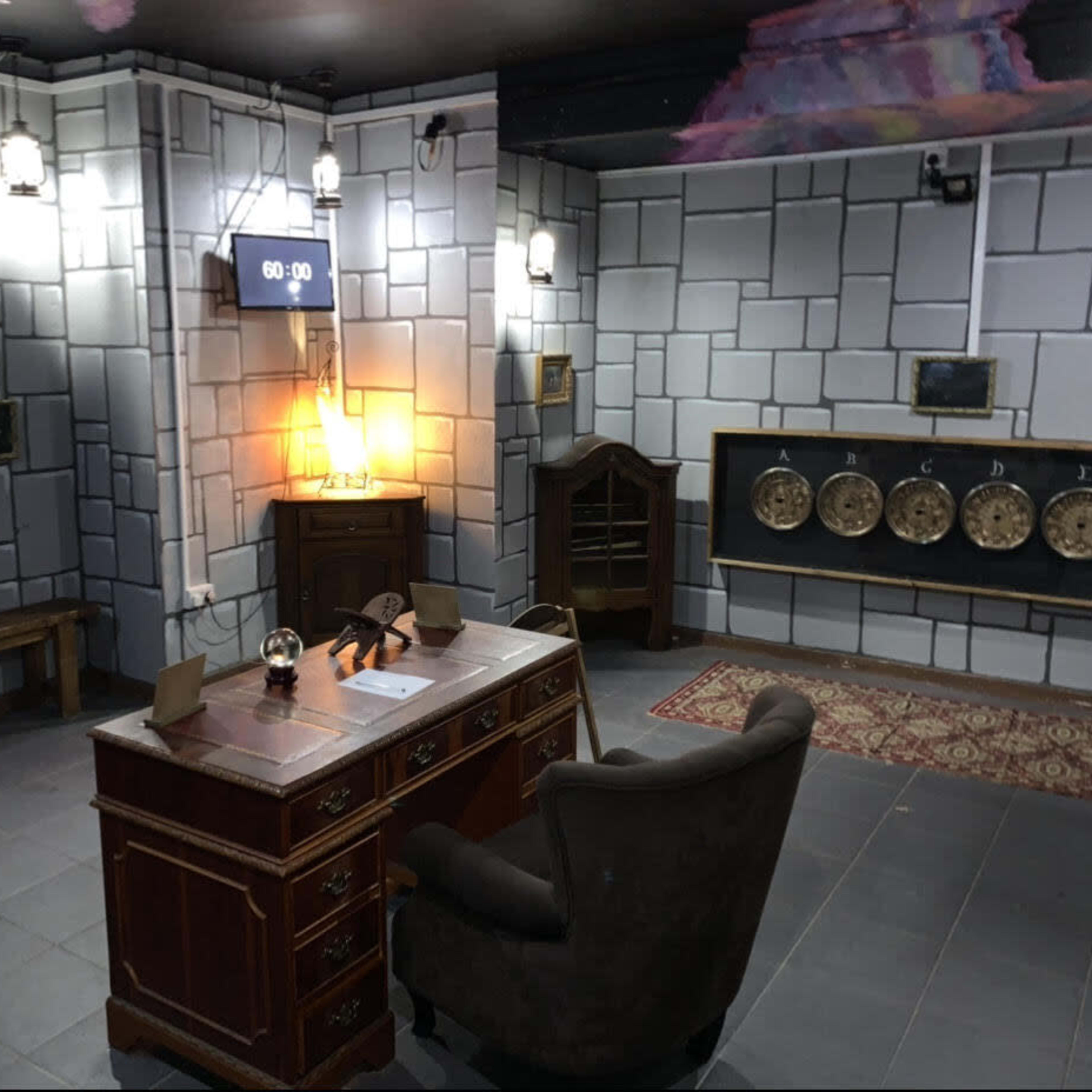 An escape room setup featuring a wooden desk with a chair, a clock displaying 60:00, and various decorative elements on the walls, including photographs and a display of circular items.