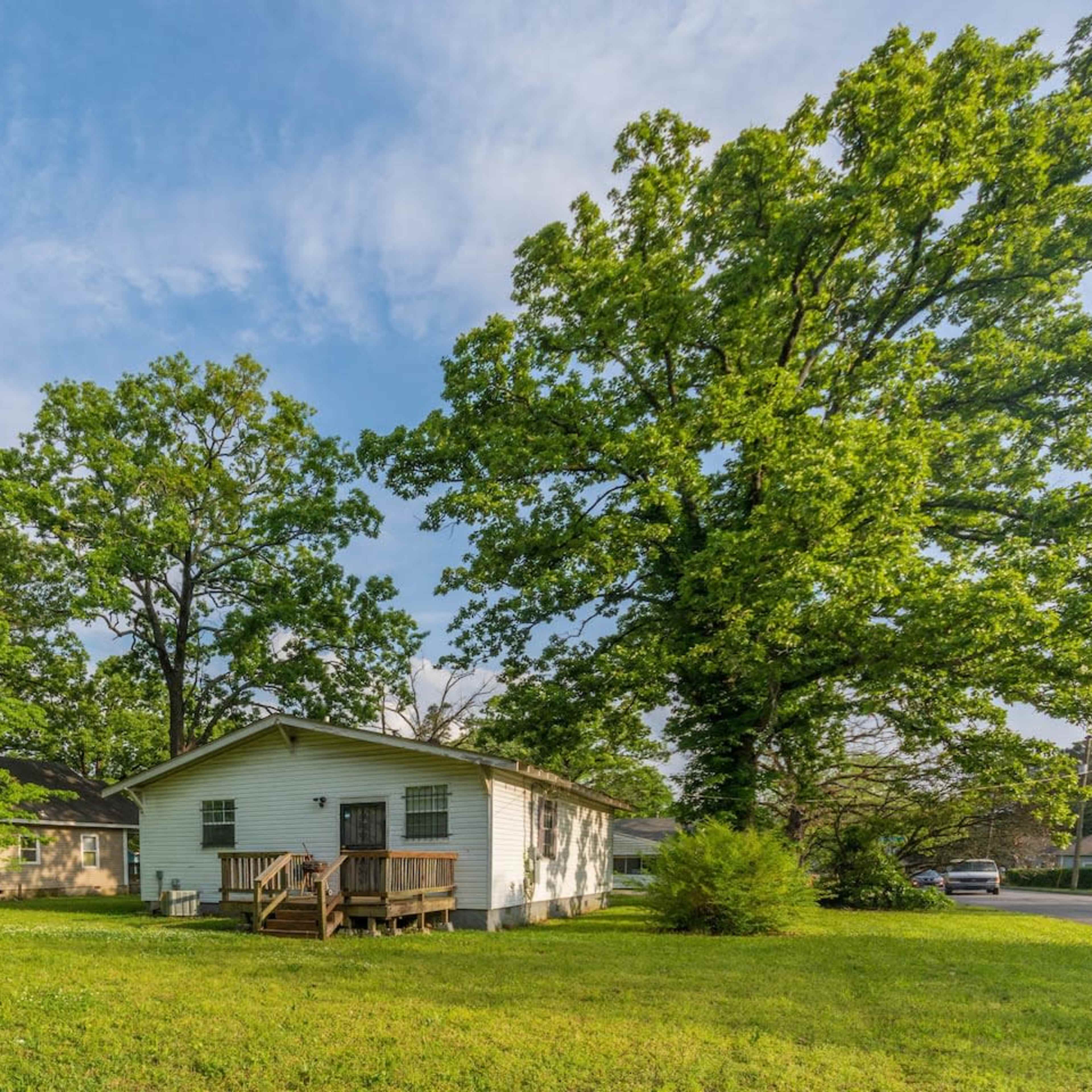 A single-story white house with a porch is surrounded by lush green trees and grass.
