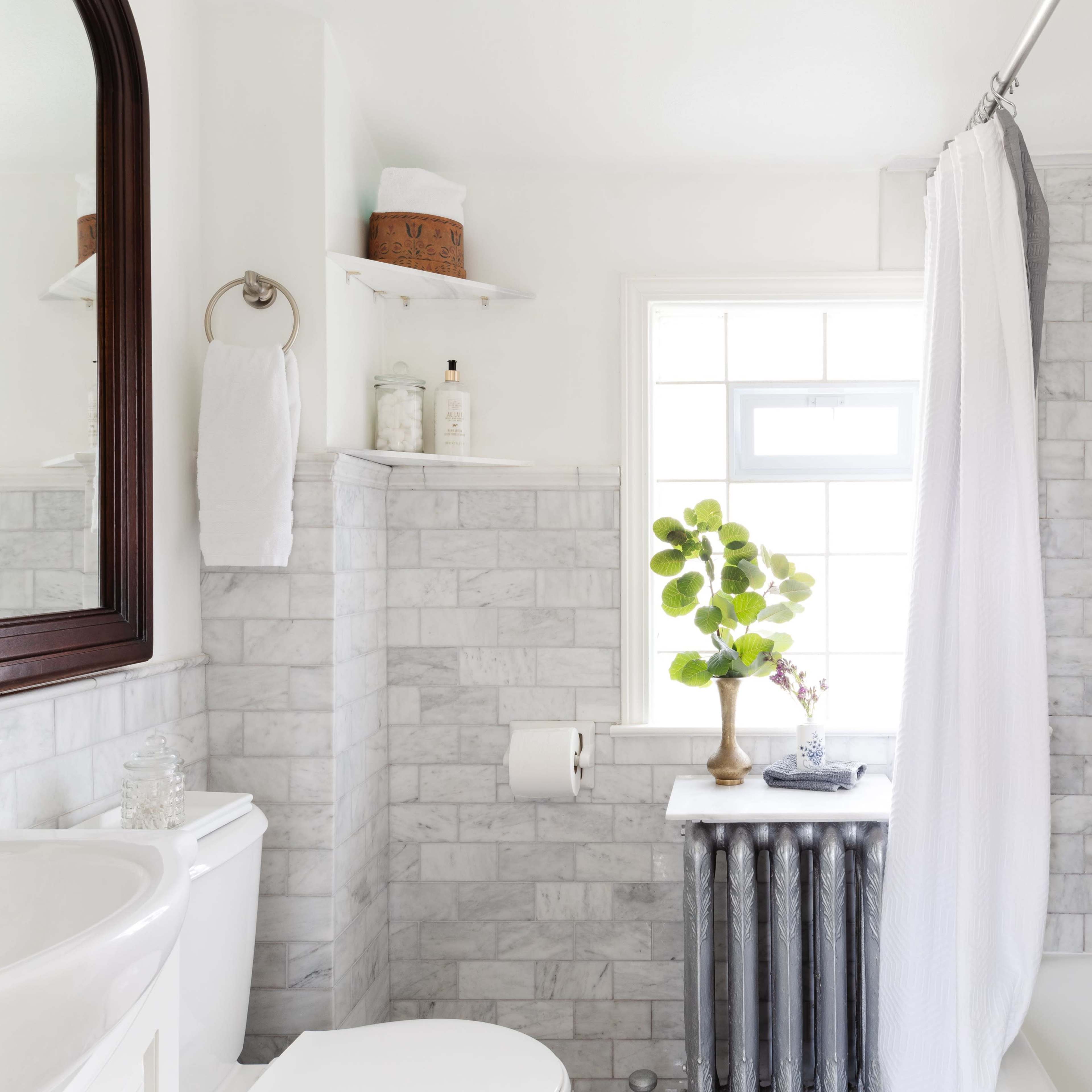 A bright bathroom features marble walls, a white shower curtain, and a small window illuminating a plant on a countertop next to a vintage radiator.