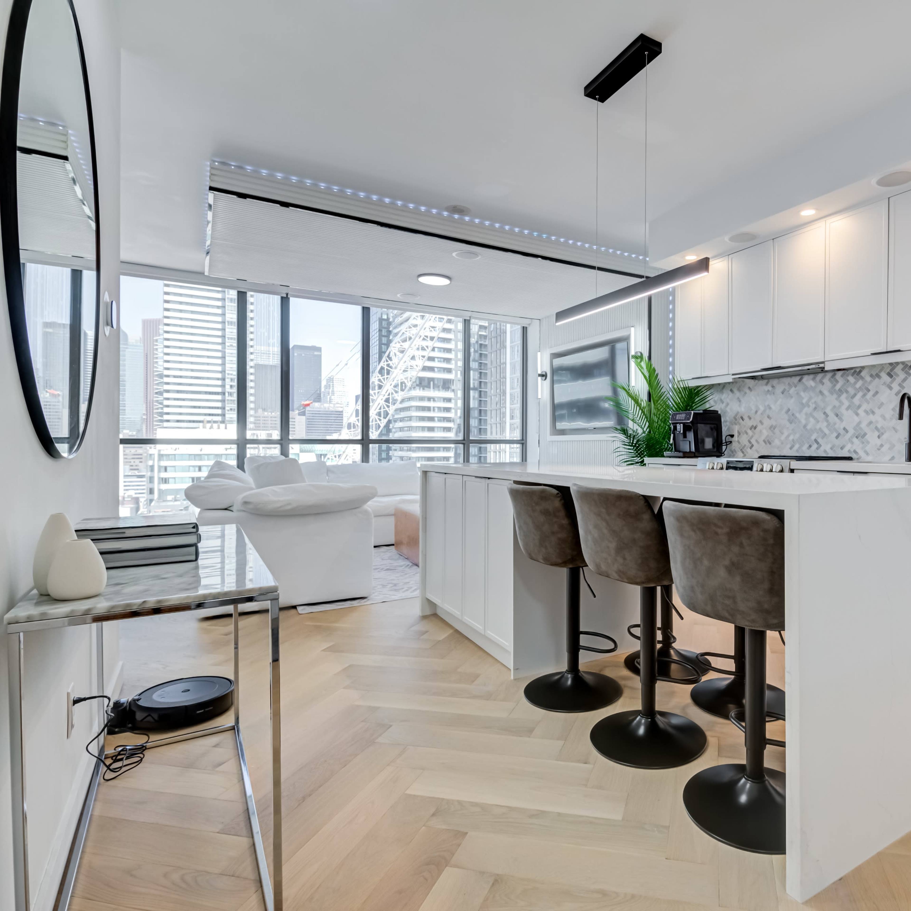 A modern kitchen features white cabinetry and a central island with four bar stools, overlooking a city skyline through large windows.