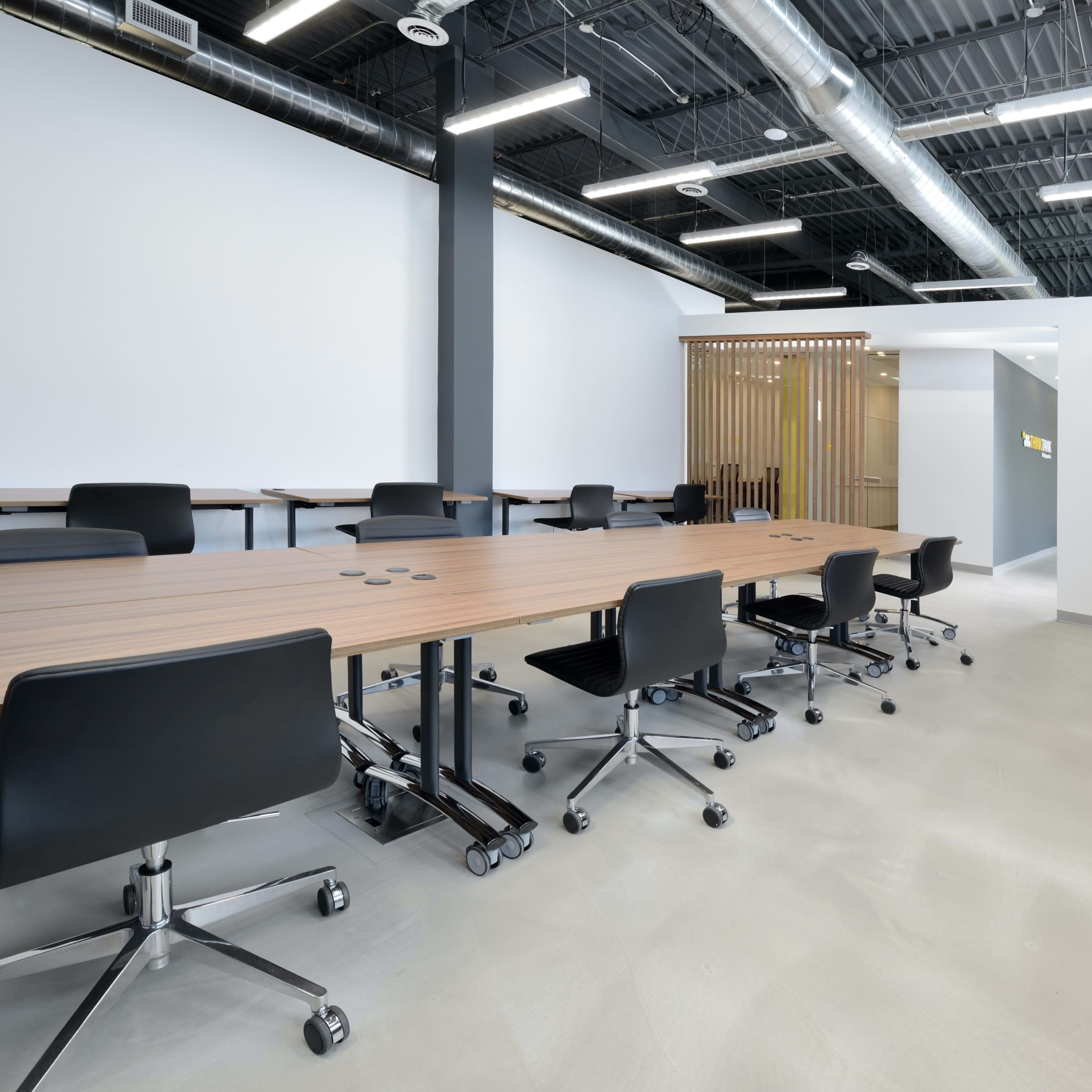 A modern conference room with long wooden tables and black swivel chairs arranged neatly.