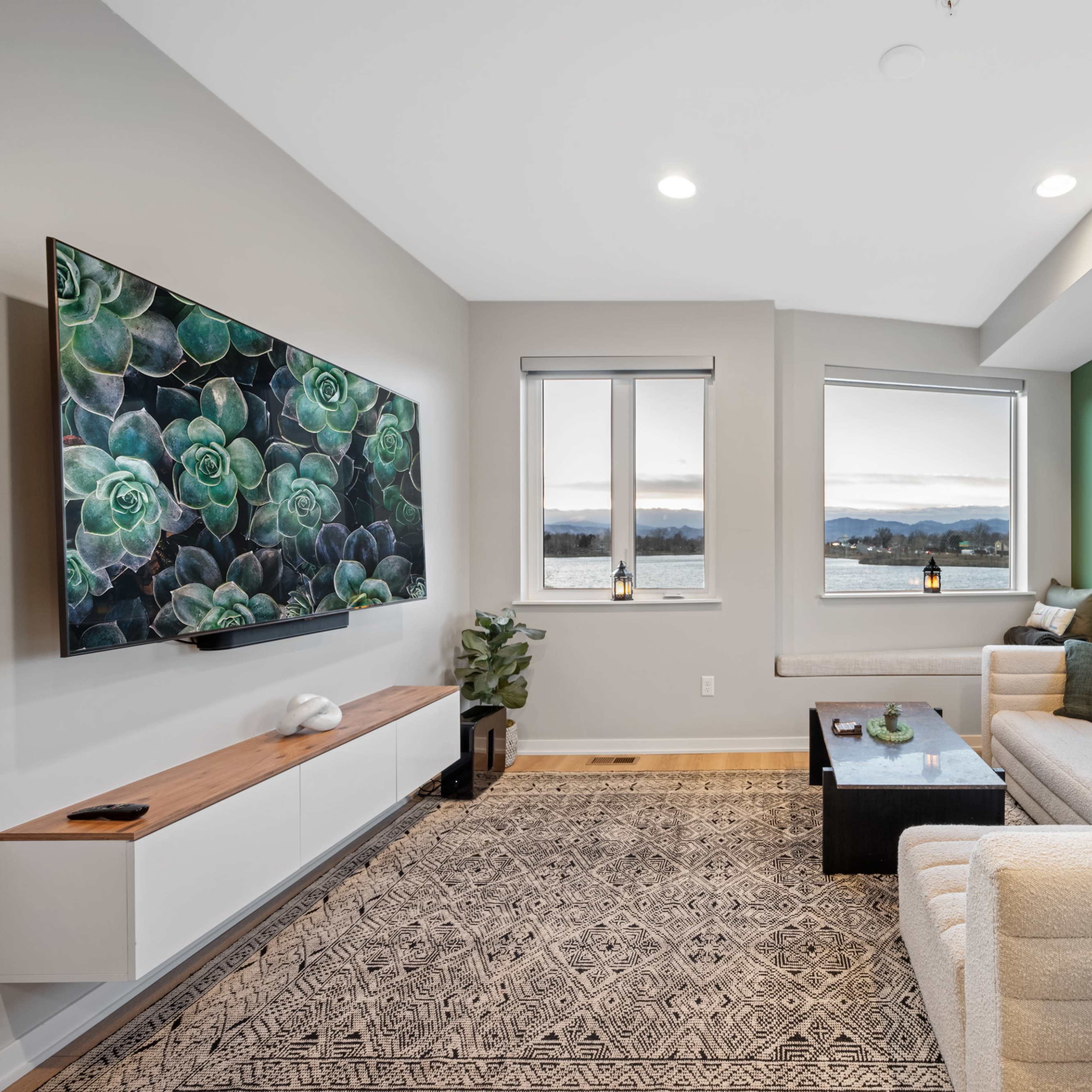 A modern living room features a large TV mounted on a wall, a beige sectional sofa, a coffee table, and windows with a view of the water.