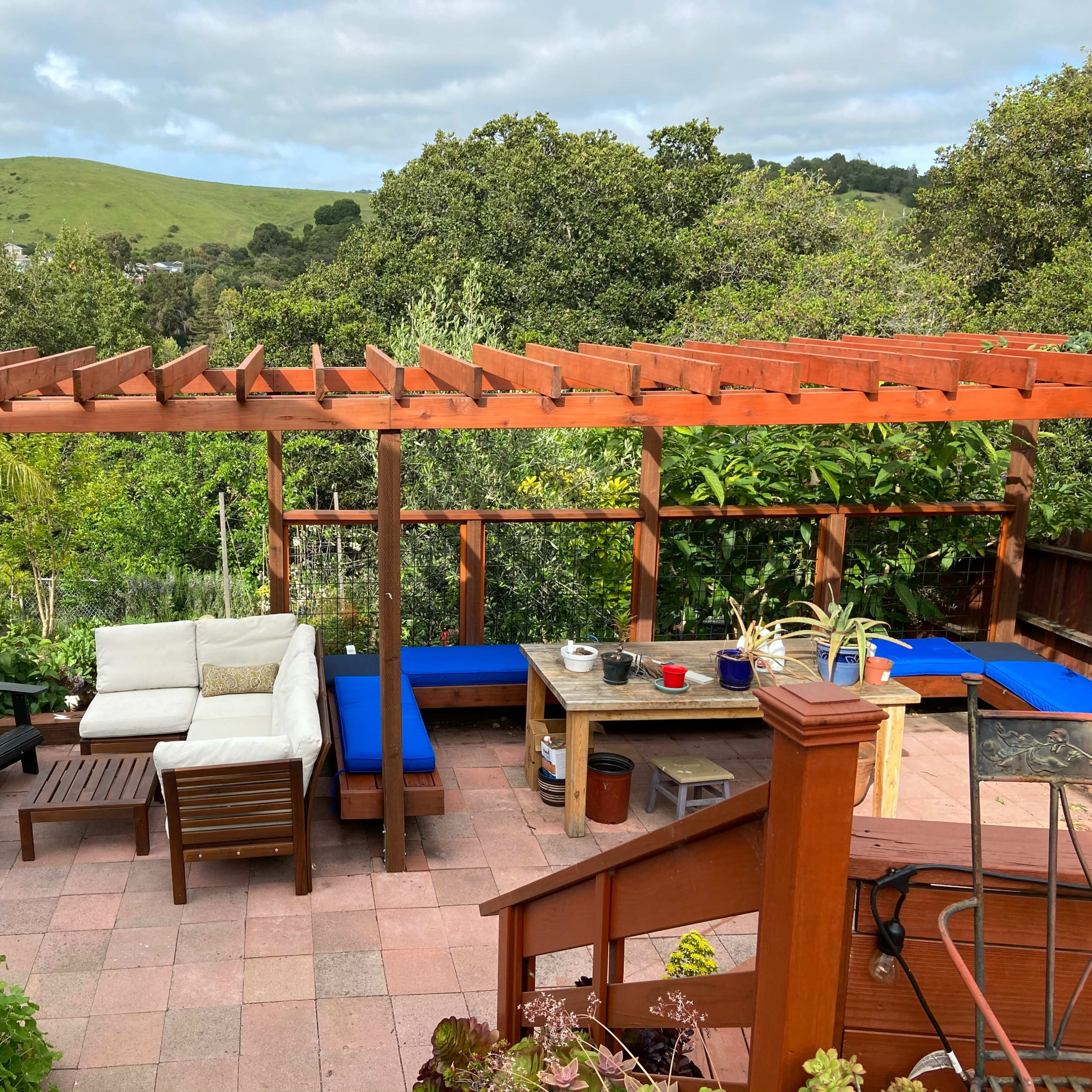 A backyard garden with a patio featuring a wooden pergola, outdoor seating, and potted plants surrounded by greenery and hills in the background.