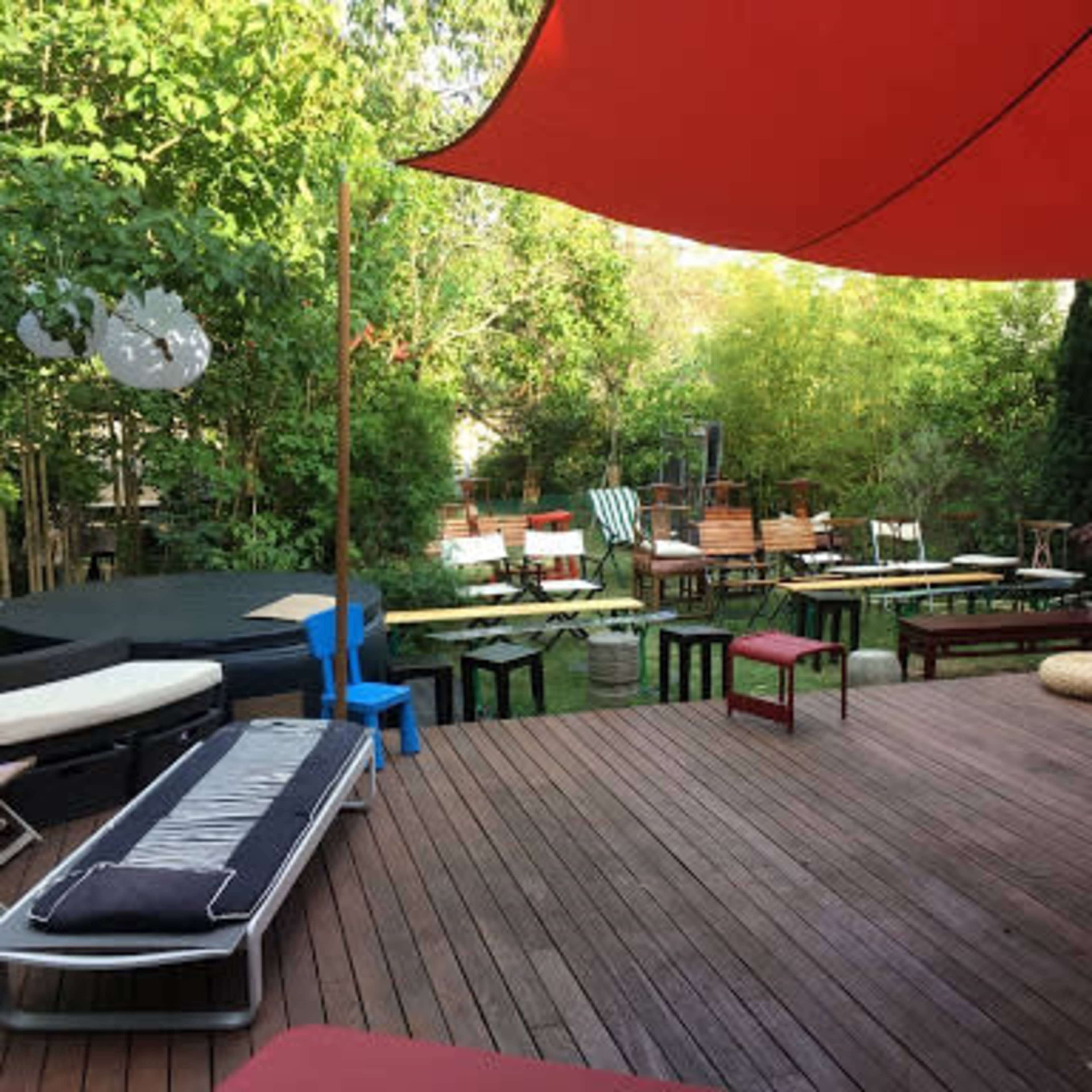 A wooden deck area in a garden with various seating arrangements, tables, and a large red canopy overhead.