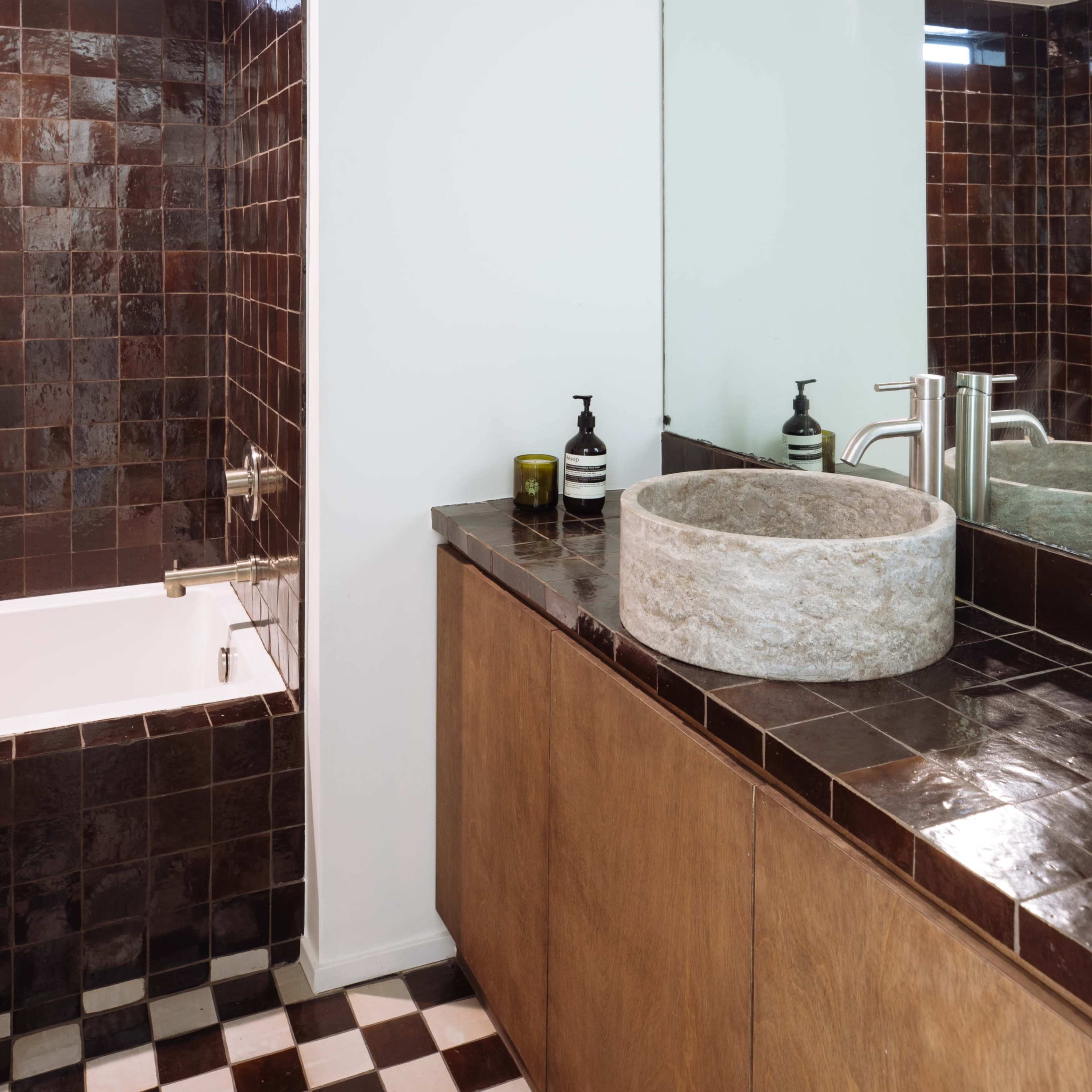 A modern bathroom with a stone sink, wooden cabinetry, dark tiled walls, and a bathtub adjacent to a shower area.