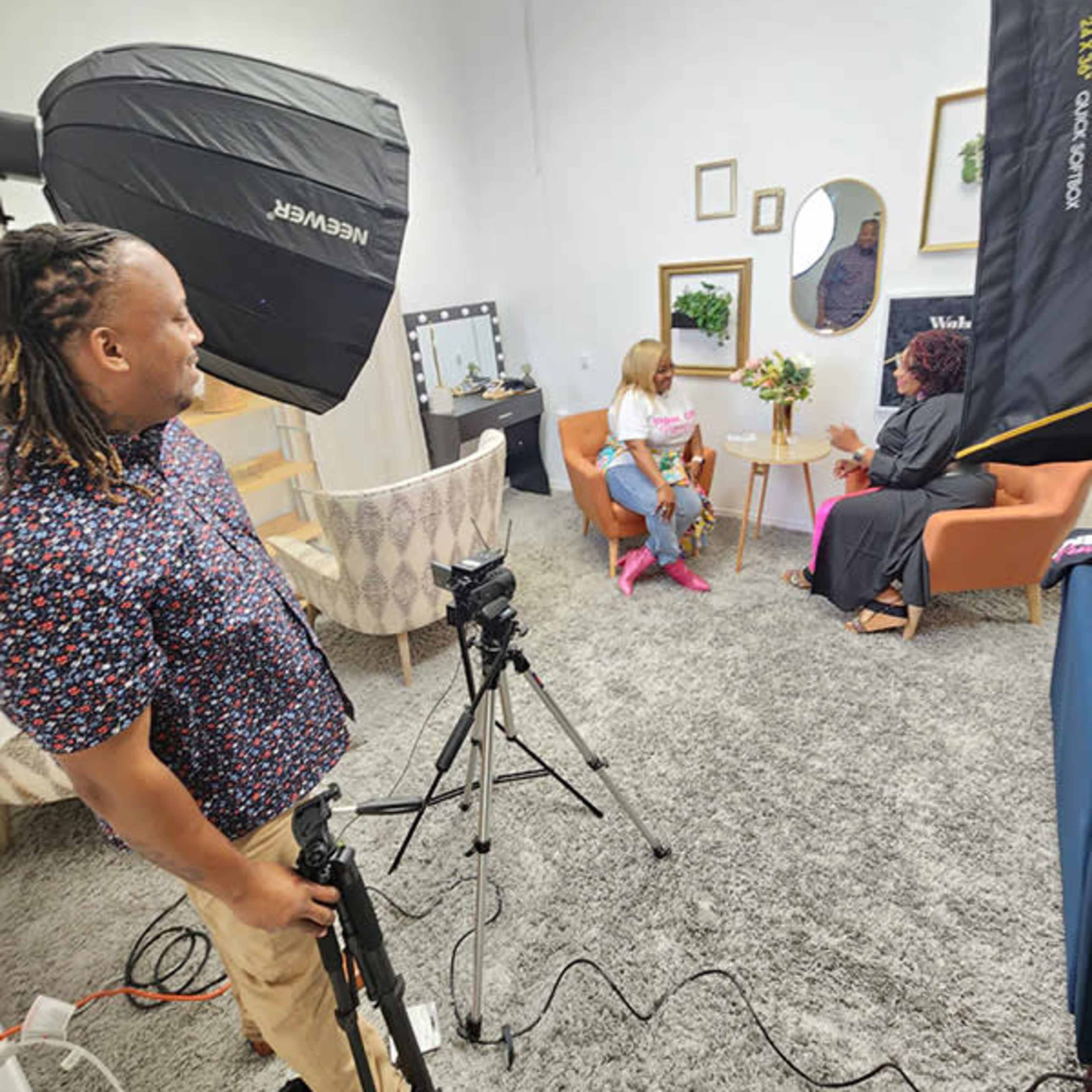 A man stands with a camera on a tripod while two women sit in an indoor setting decorated with plants and framed artwork.