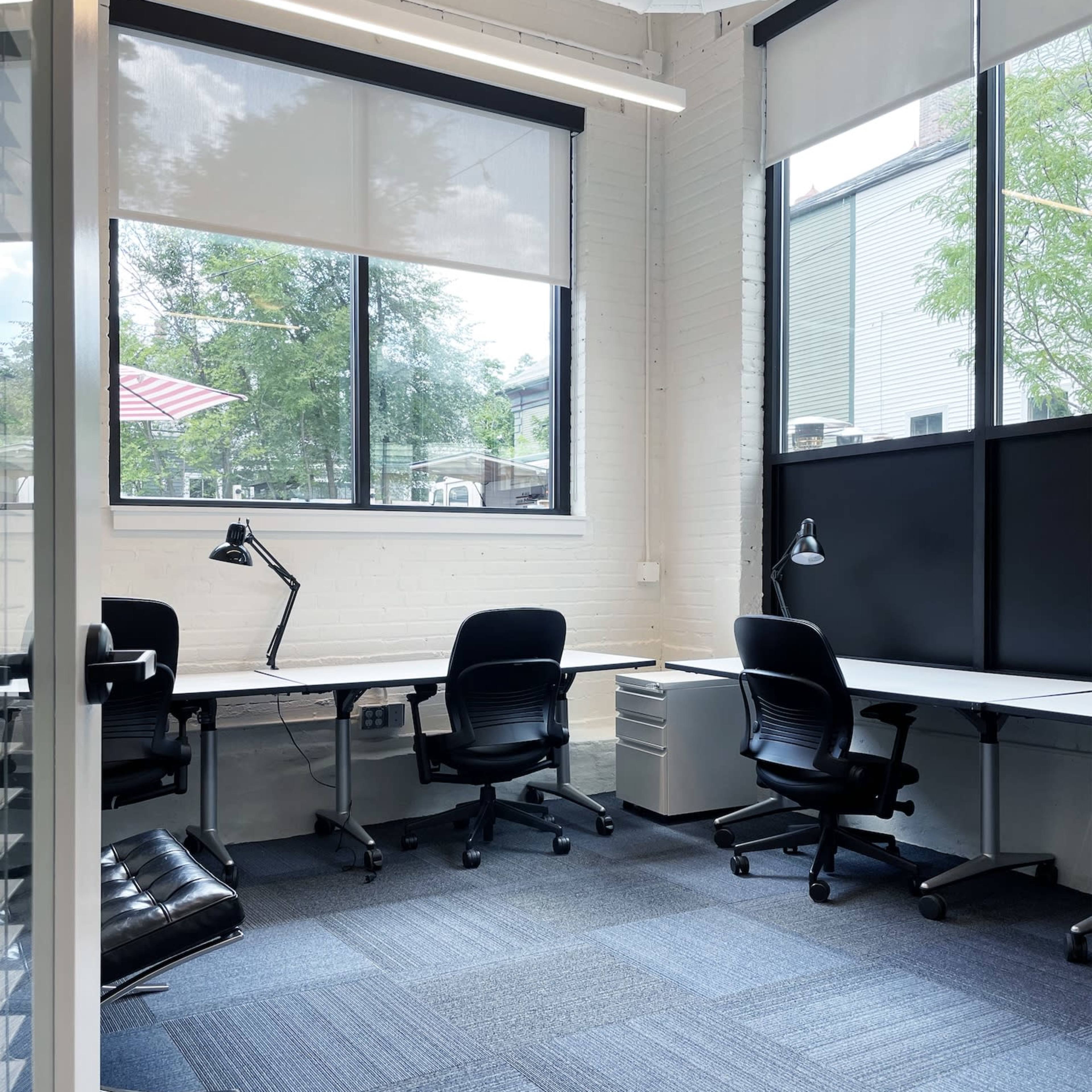 A modern office space featuring two desks, office chairs, and large windows with natural light.