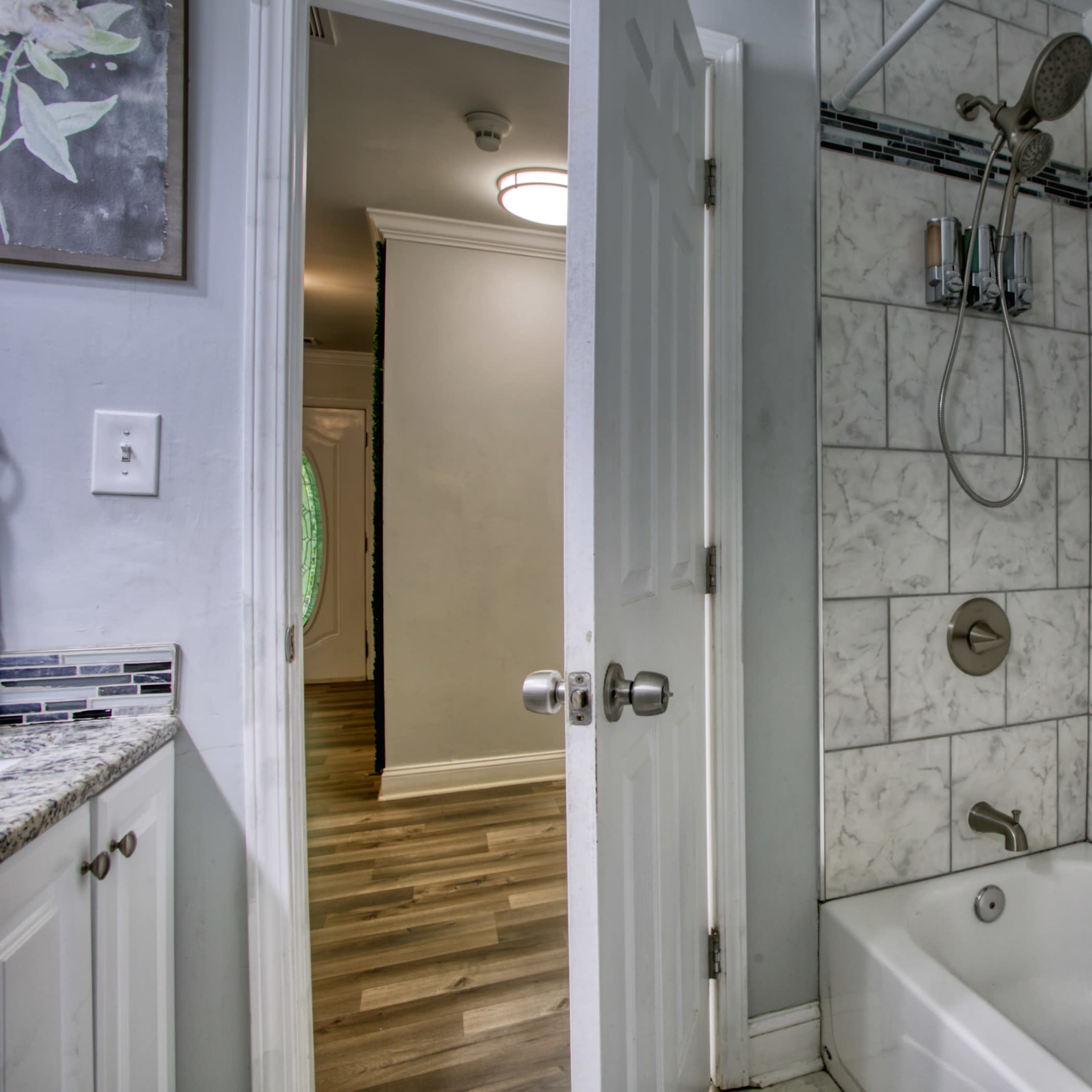 A bathroom with a white tub and showerhead, featuring tiled walls and an open door leading to a hallway with wooden flooring.