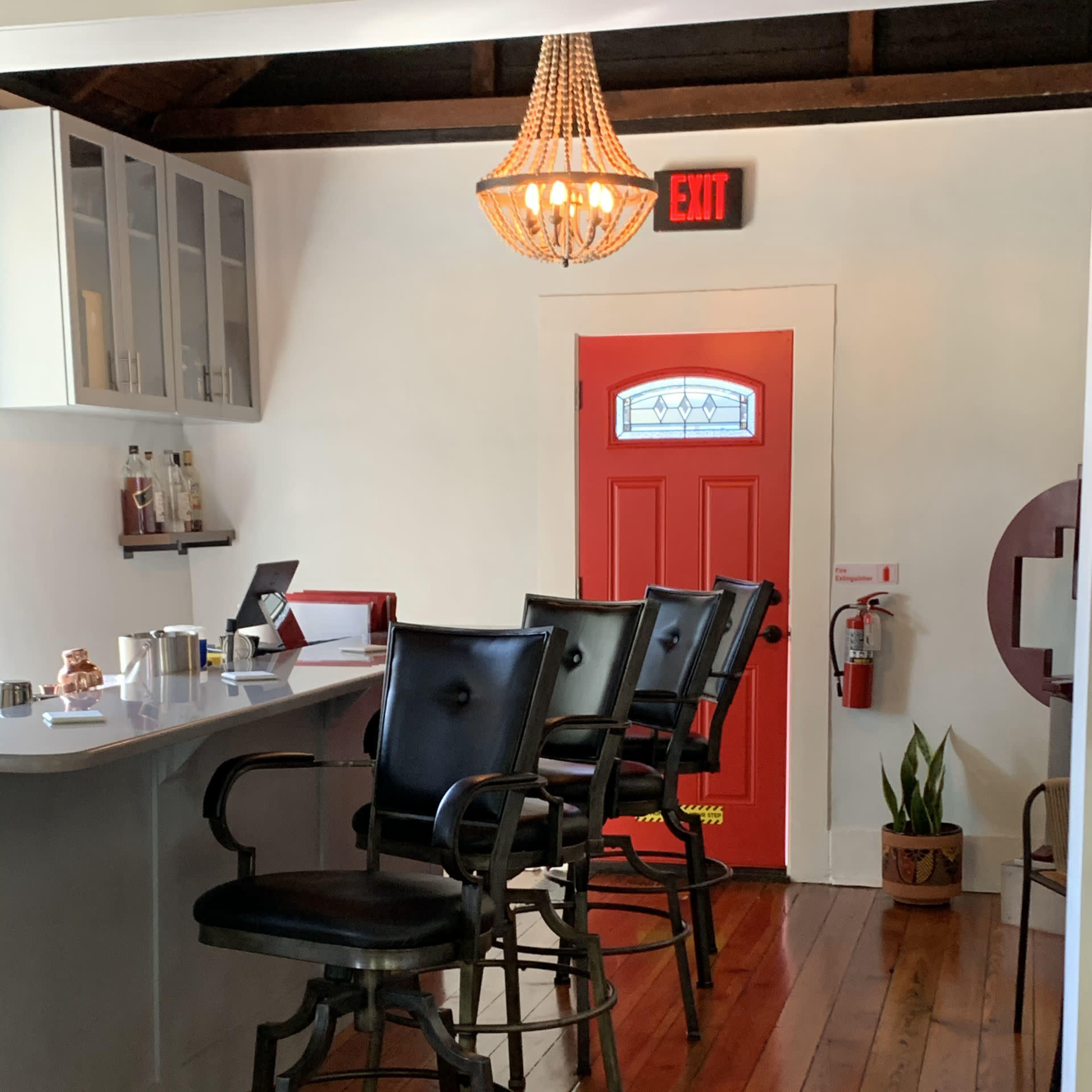 A well-lit room with high black bar stools positioned in front of a counter, featuring a vibrant red door and a chandelier overhead.