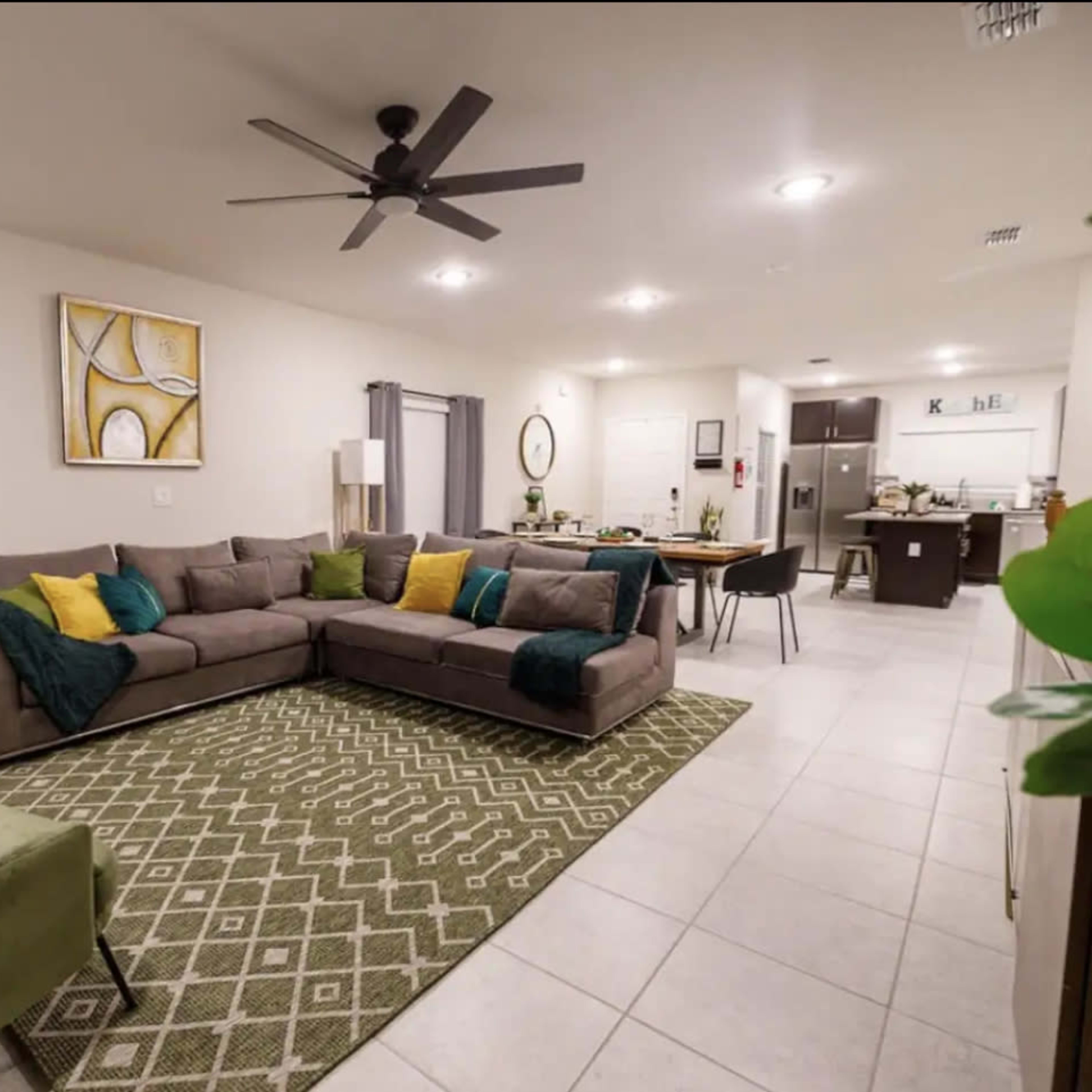 A spacious living area features a large gray sectional sofa with green and yellow cushions, a patterned rug, and a connected dining space with a kitchen in the background.