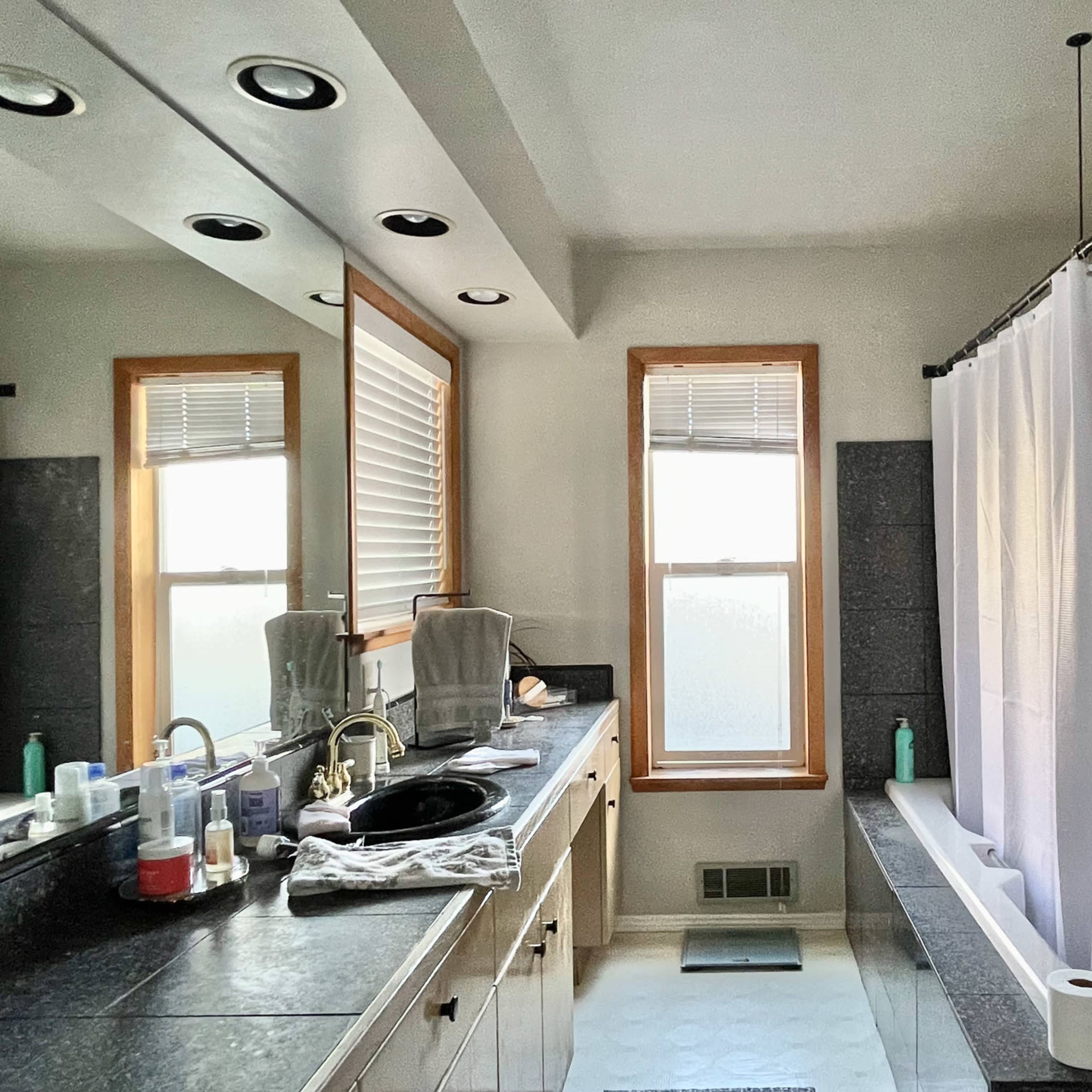 A bathroom featuring a shower curtain, sink with toiletries, and two windows with white blinds.
