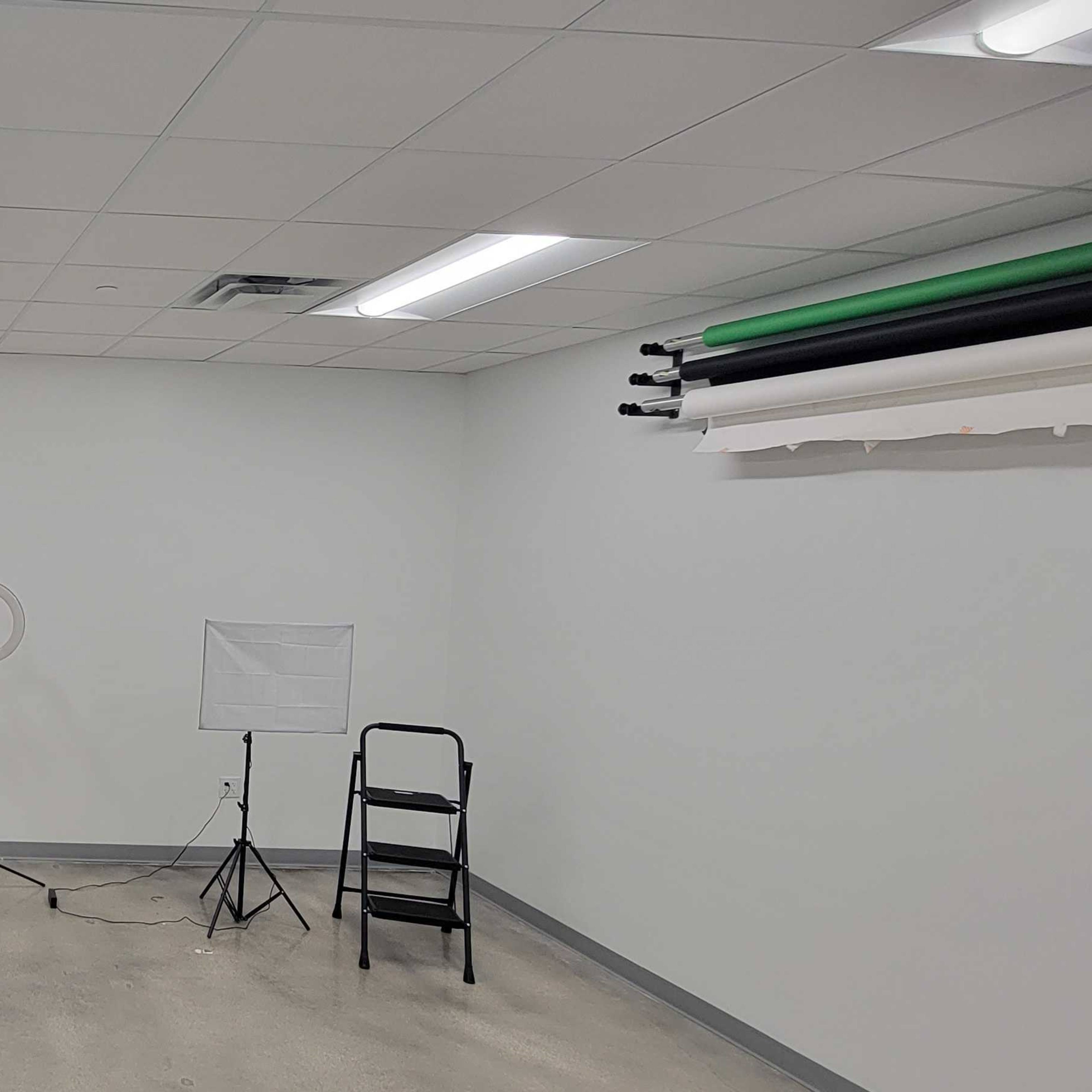 A light-colored room with a few pieces of furniture, including a table, a mirror, a ring light, and a folding chair, along with rolls of backdrop paper on the wall.