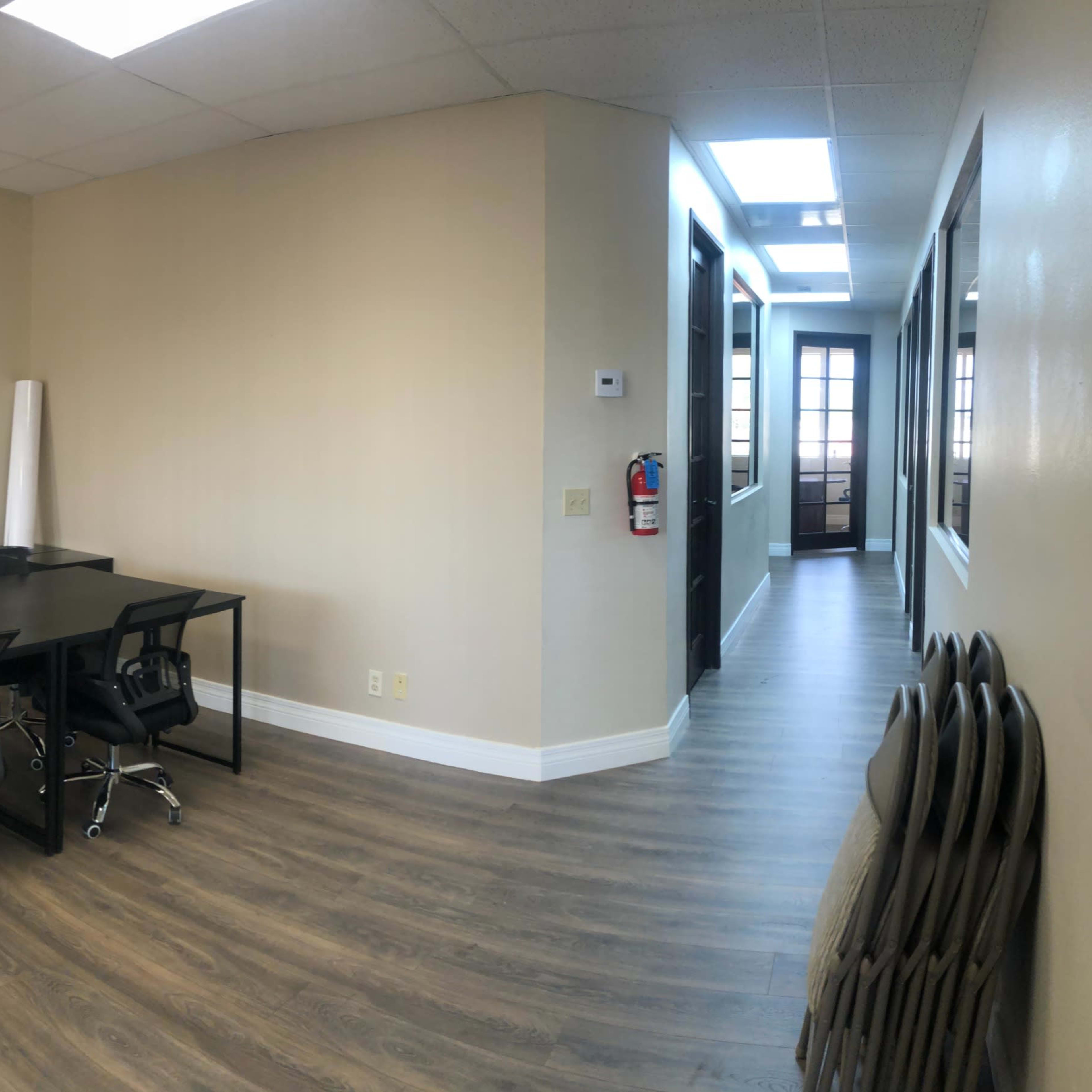 A modern office space featuring a conference table with chairs, light-colored walls, and wooden flooring, along with a hallway leading to additional rooms.