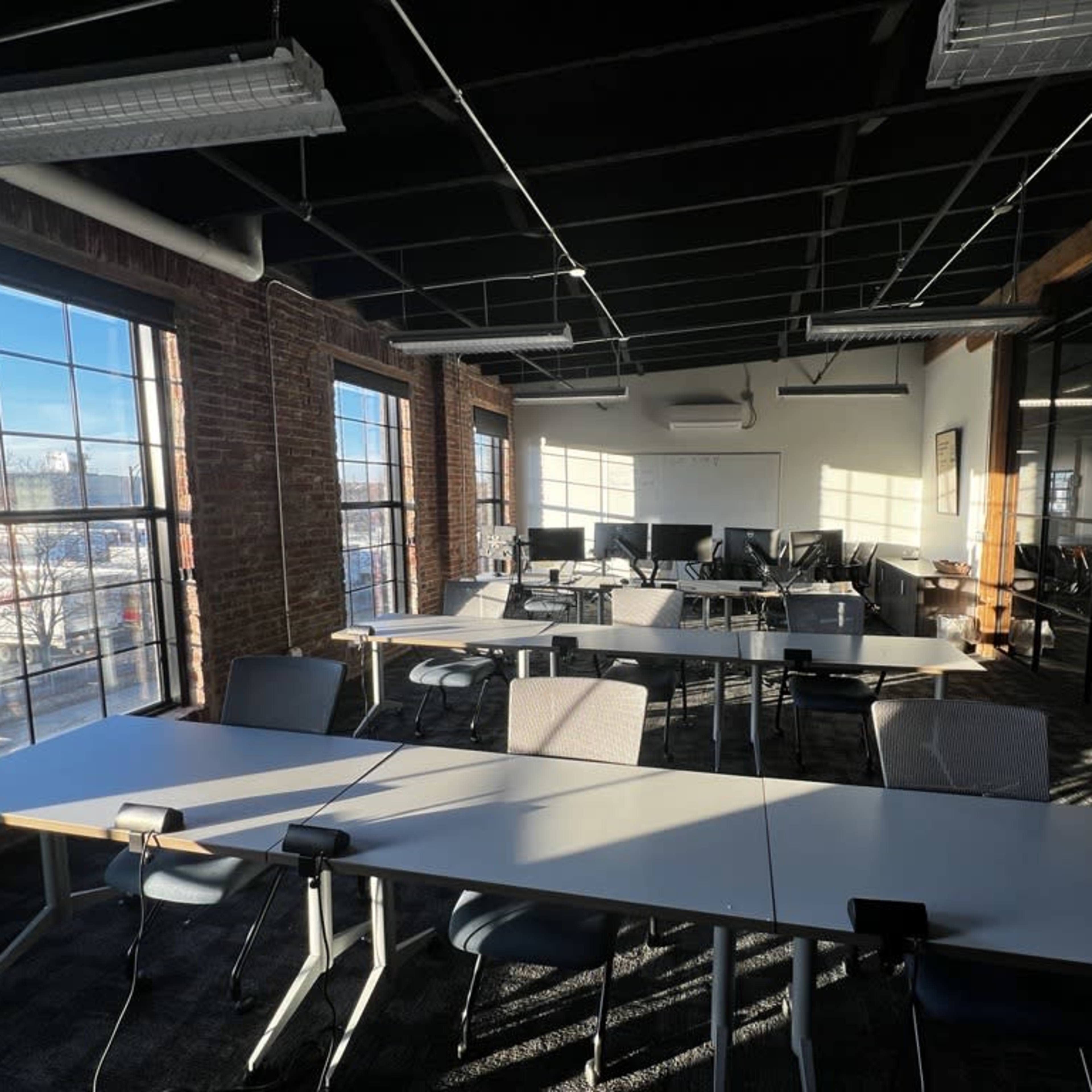 A bright, modern classroom with rows of tables and chairs arranged in a spacious area featuring large windows and exposed brick walls.